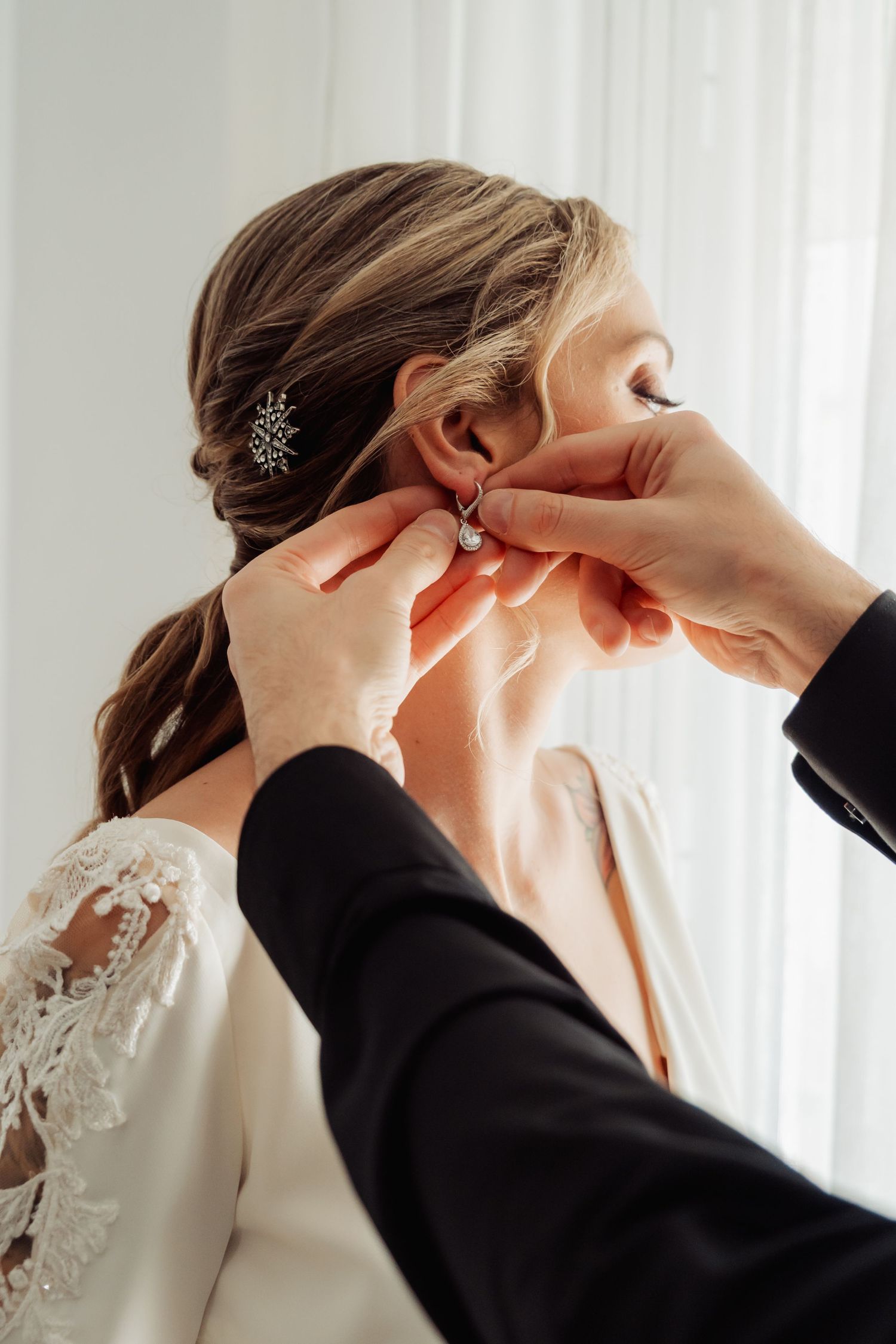 Hands carefully fastening a crystal drop earring on a bride with lace wedding dress.