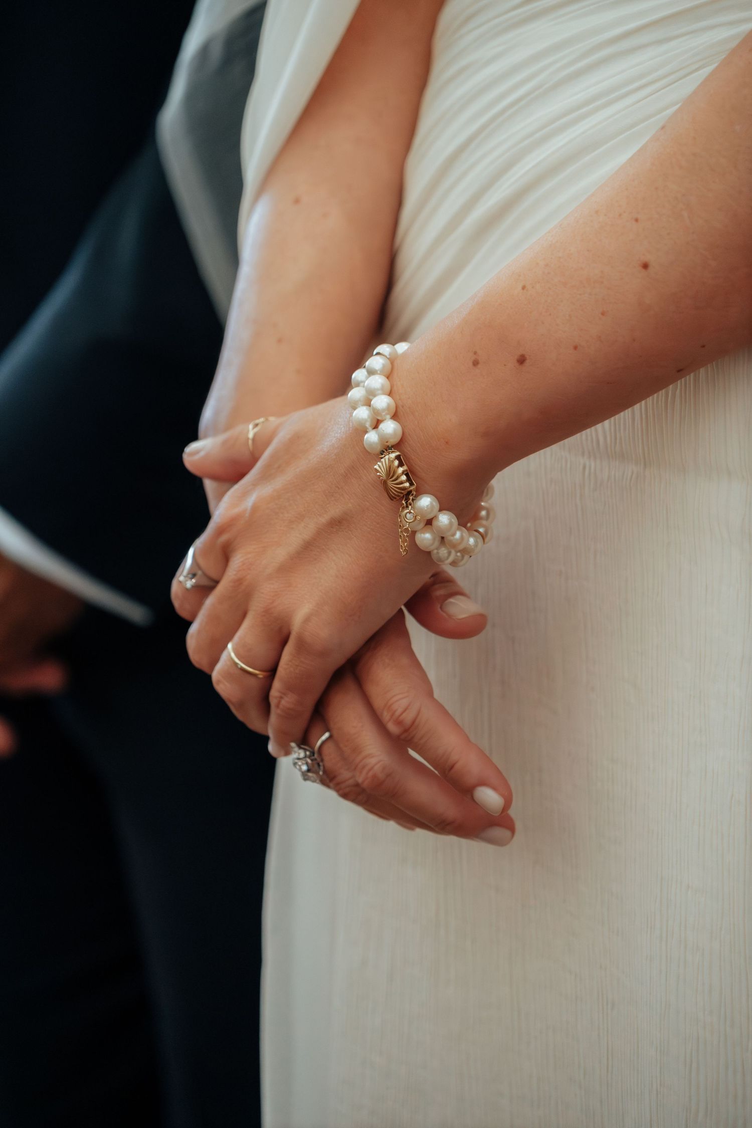 Close-up of bride's hands adorned with pearl bracelet and diamond engagement ring in white gown.