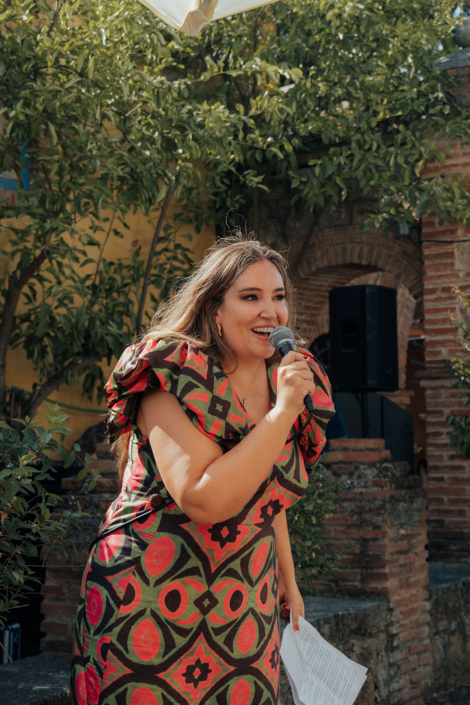 Woman in colorful patterned dress speaking into a microphone at an outdoor wedding ceremony in a garden.
