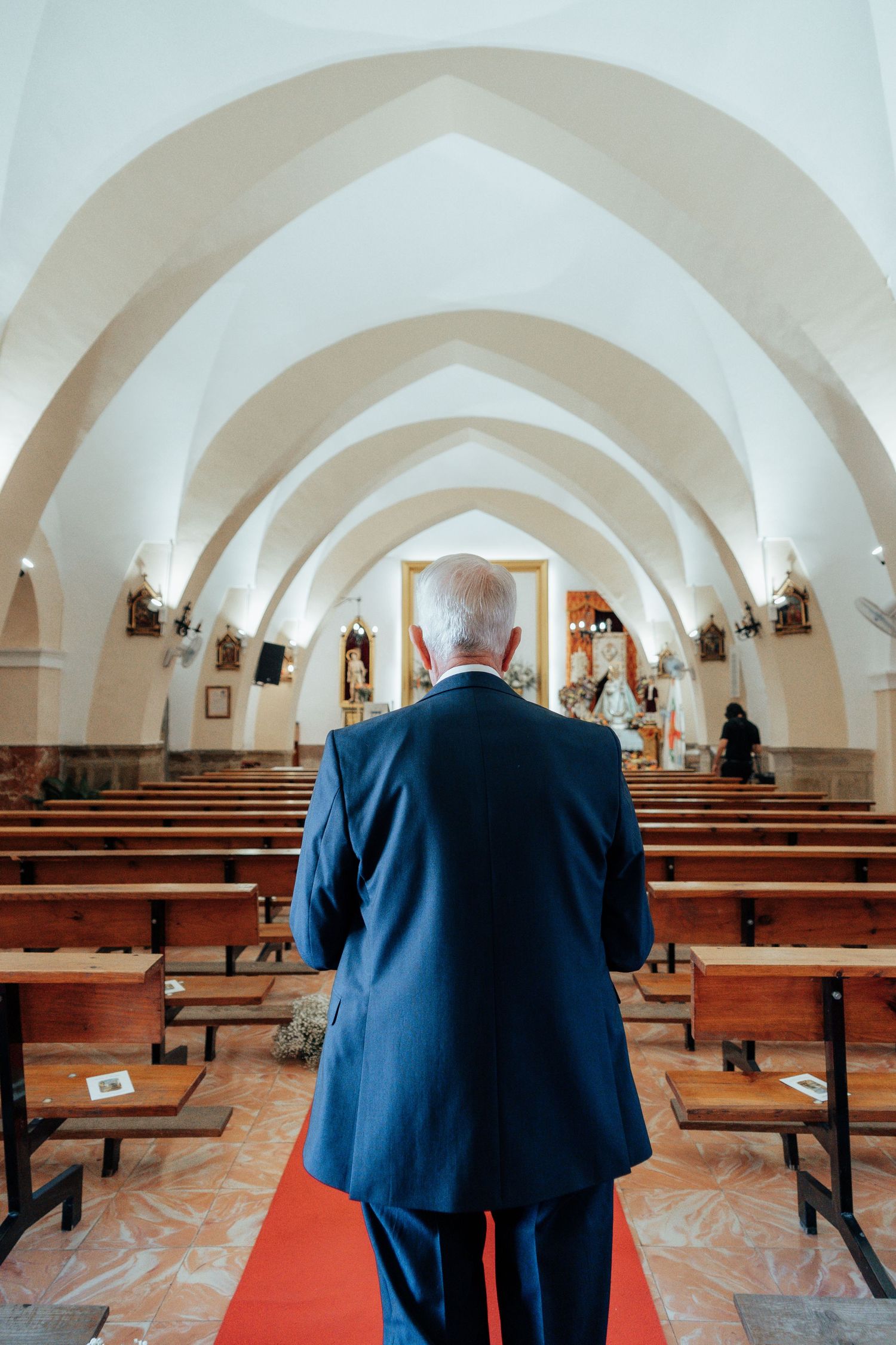 Man in blue suit walking down red carpet aisle of elegant white arched Spanish church interior.