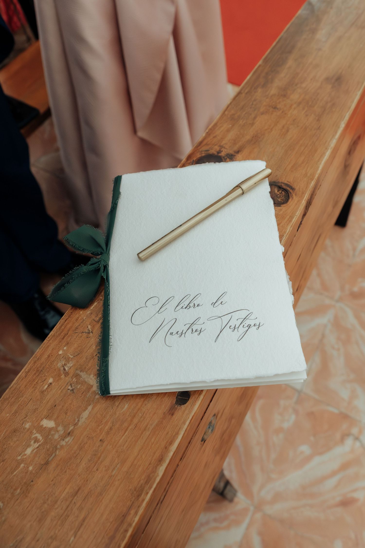 Wedding guest book titled El Libro de Nuestros Testigos with gold pen on wooden bench.