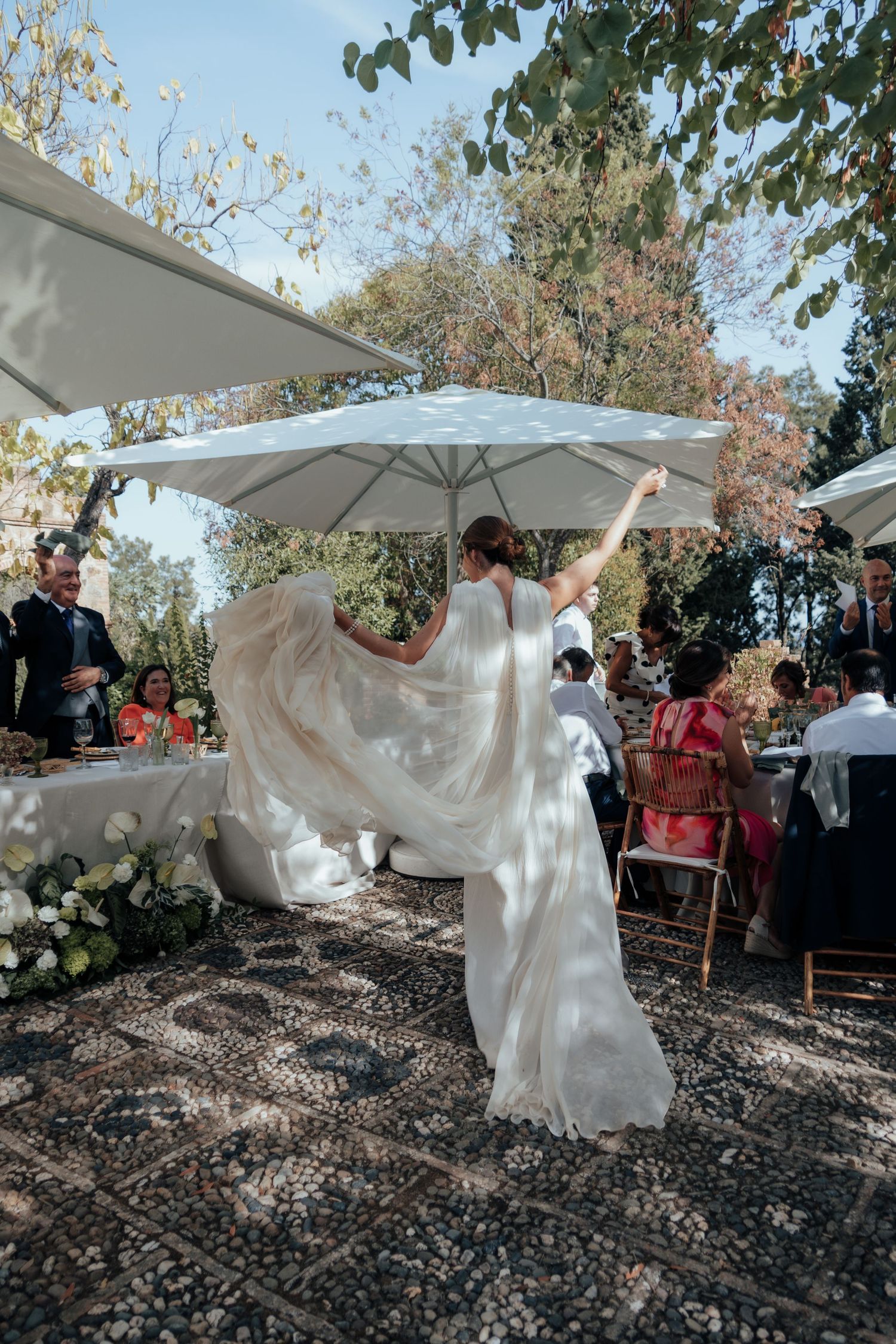 Bride twirling her flowing white veil under a white umbrella at an outdoor garden wedding reception.