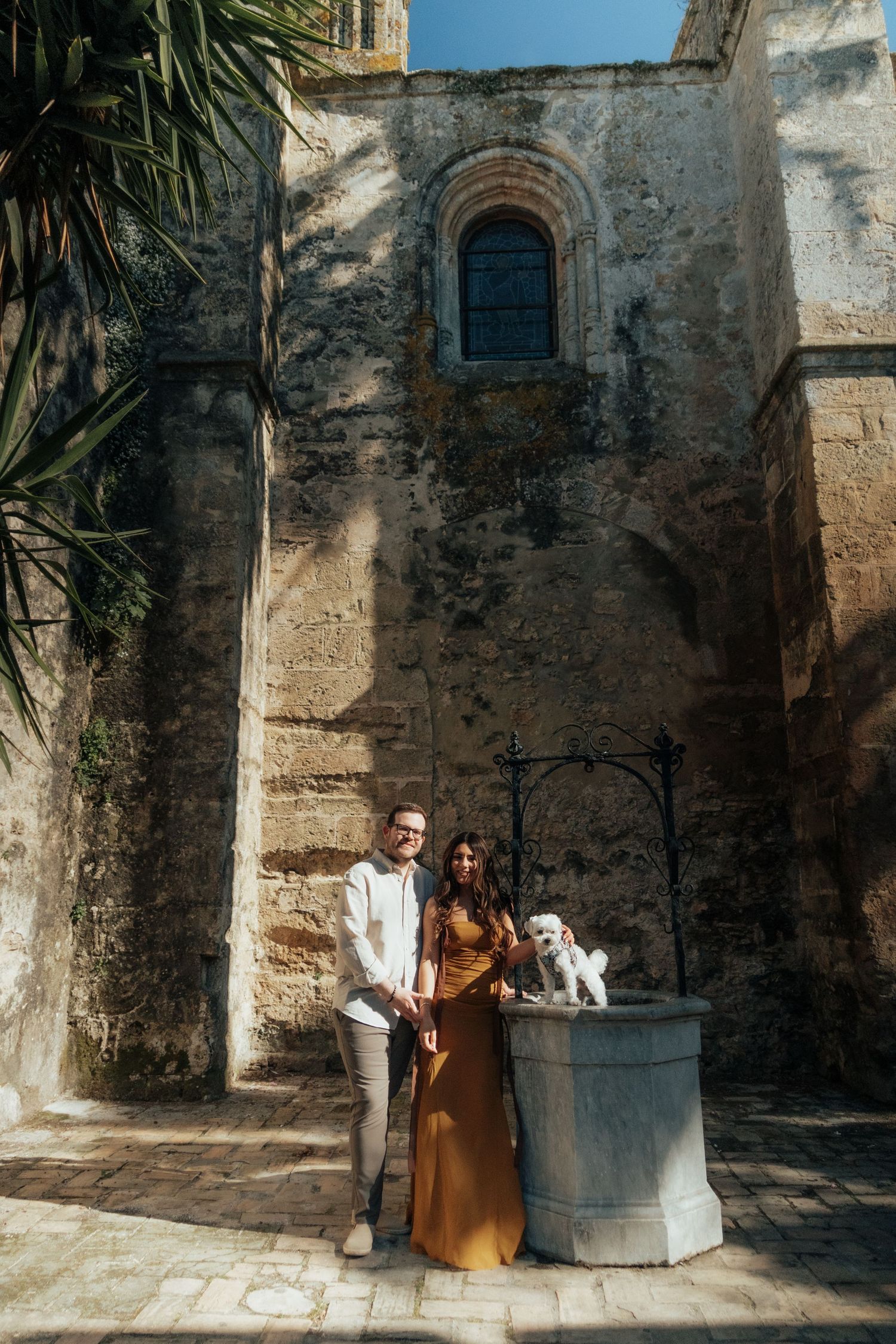 Couple in elegant attire posing by a stone well outside an ancient Mediterranean building with arched windows.