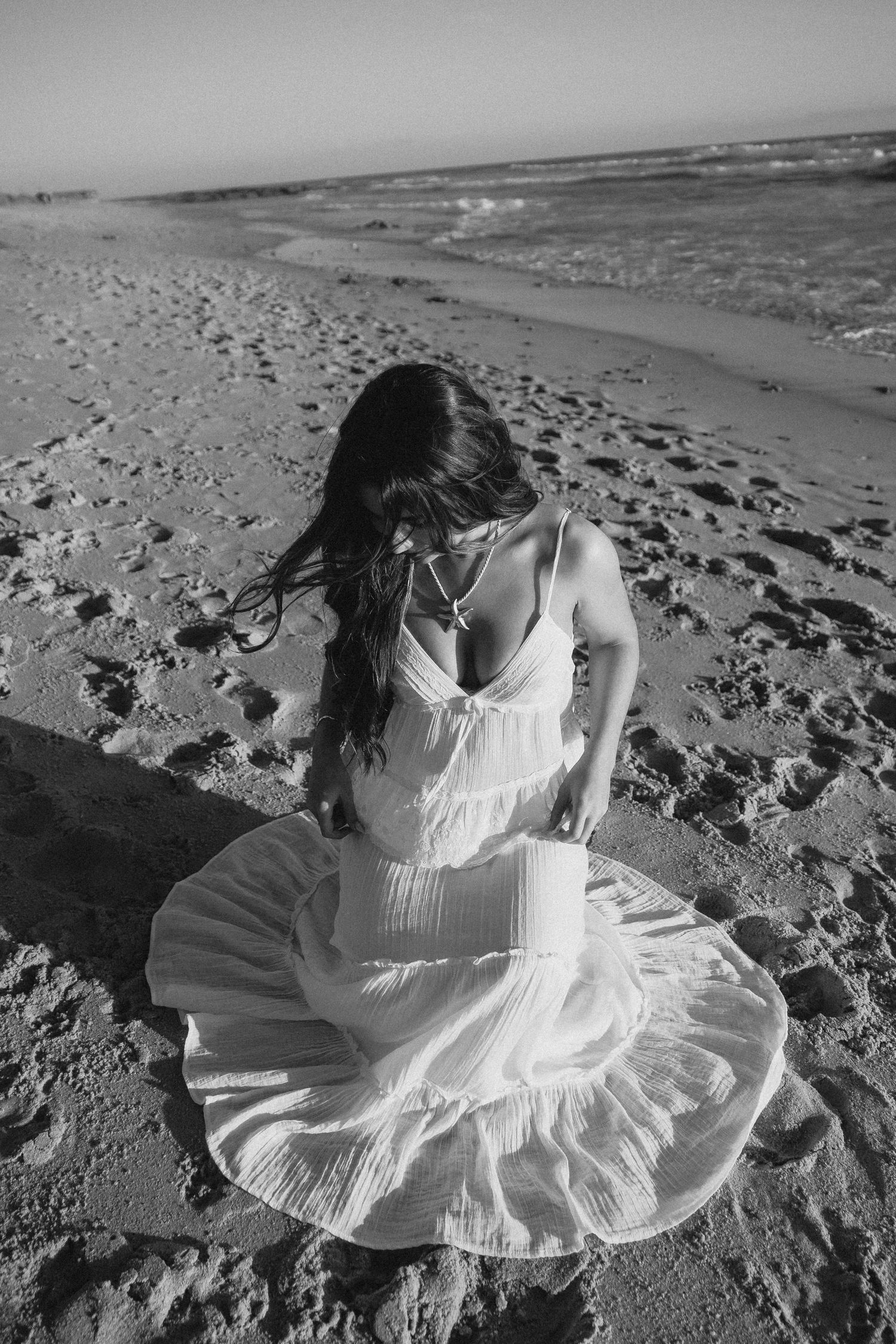 Black and white photo of a woman in a white dress sitting on a sandy beach with ocean waves in the background.