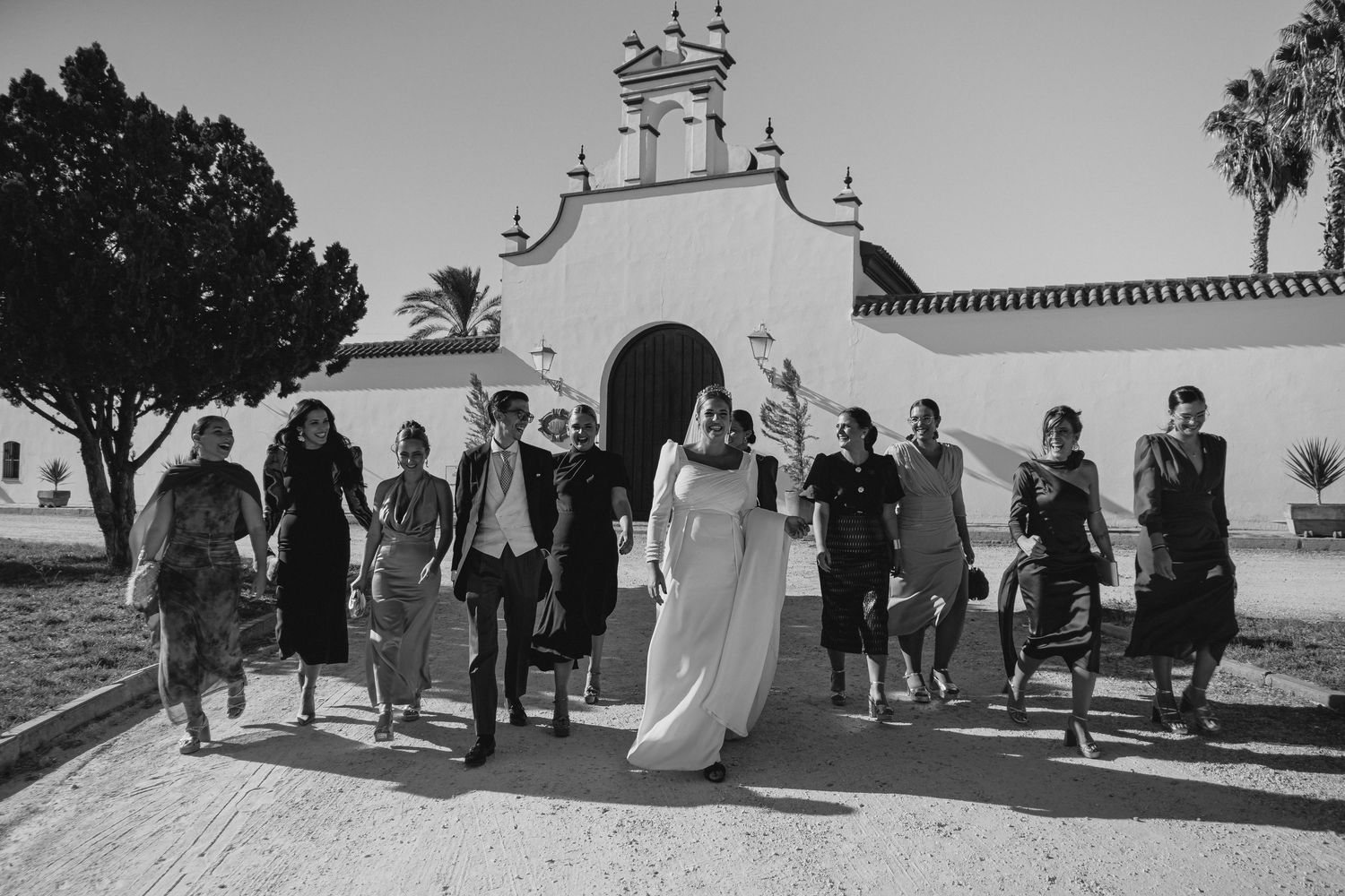 Bride in white gown walks with wedding party in front of a white Spanish-style chapel in black and white.