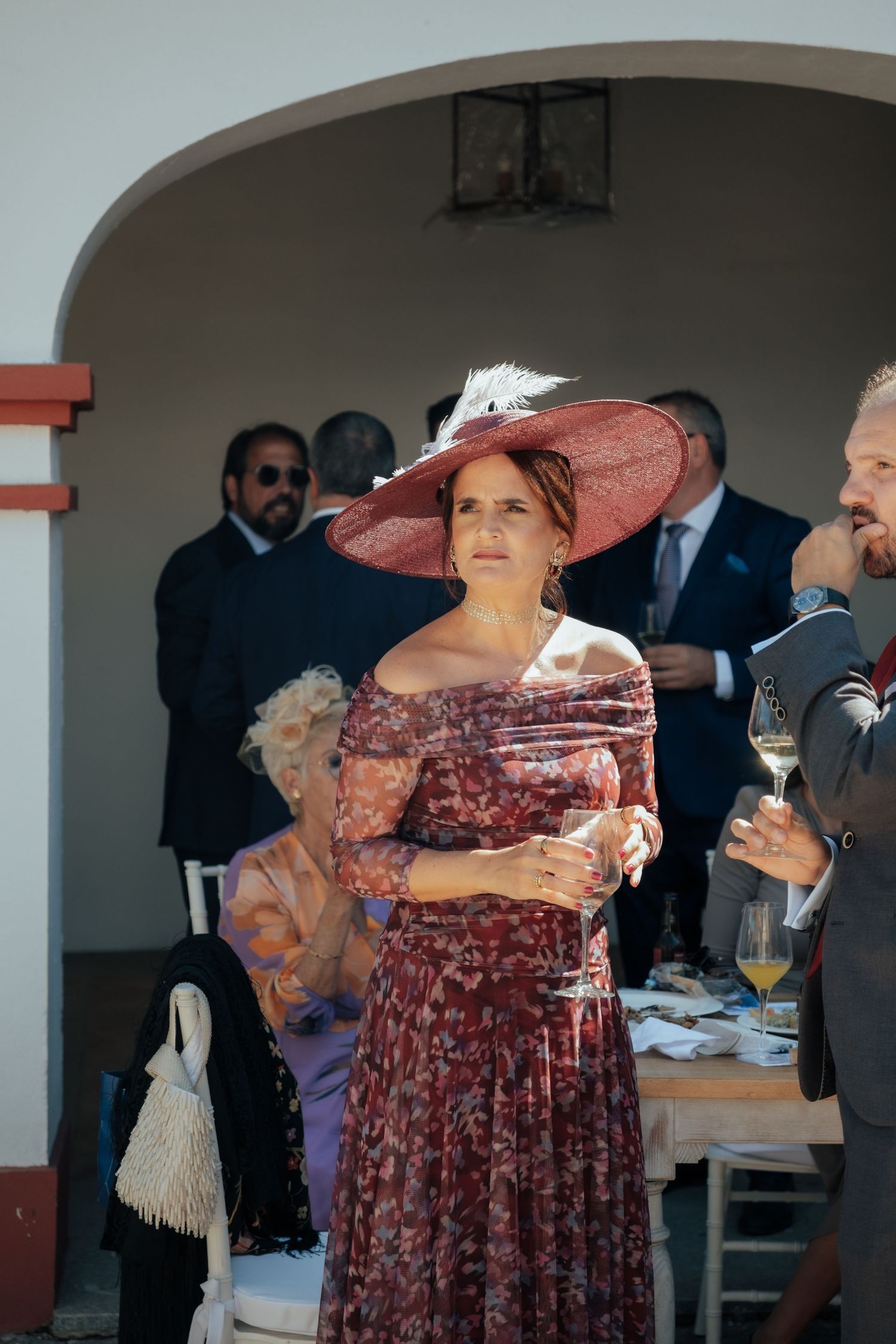 Elegant woman in floral dress and pink fascinator hat holding champagne at wedding.