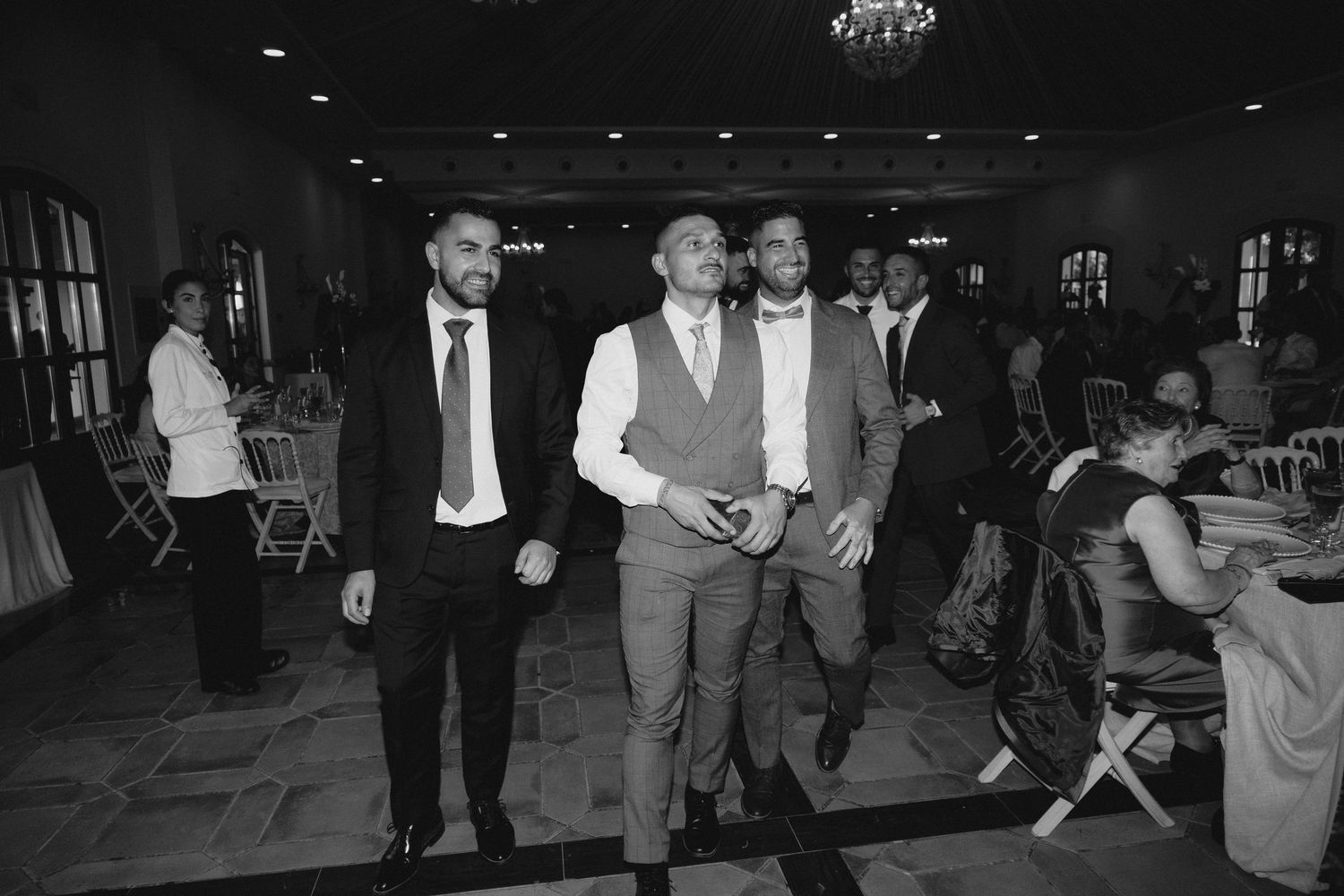 Black and white photo of groomsmen in suits walking confidently through wedding reception hall.