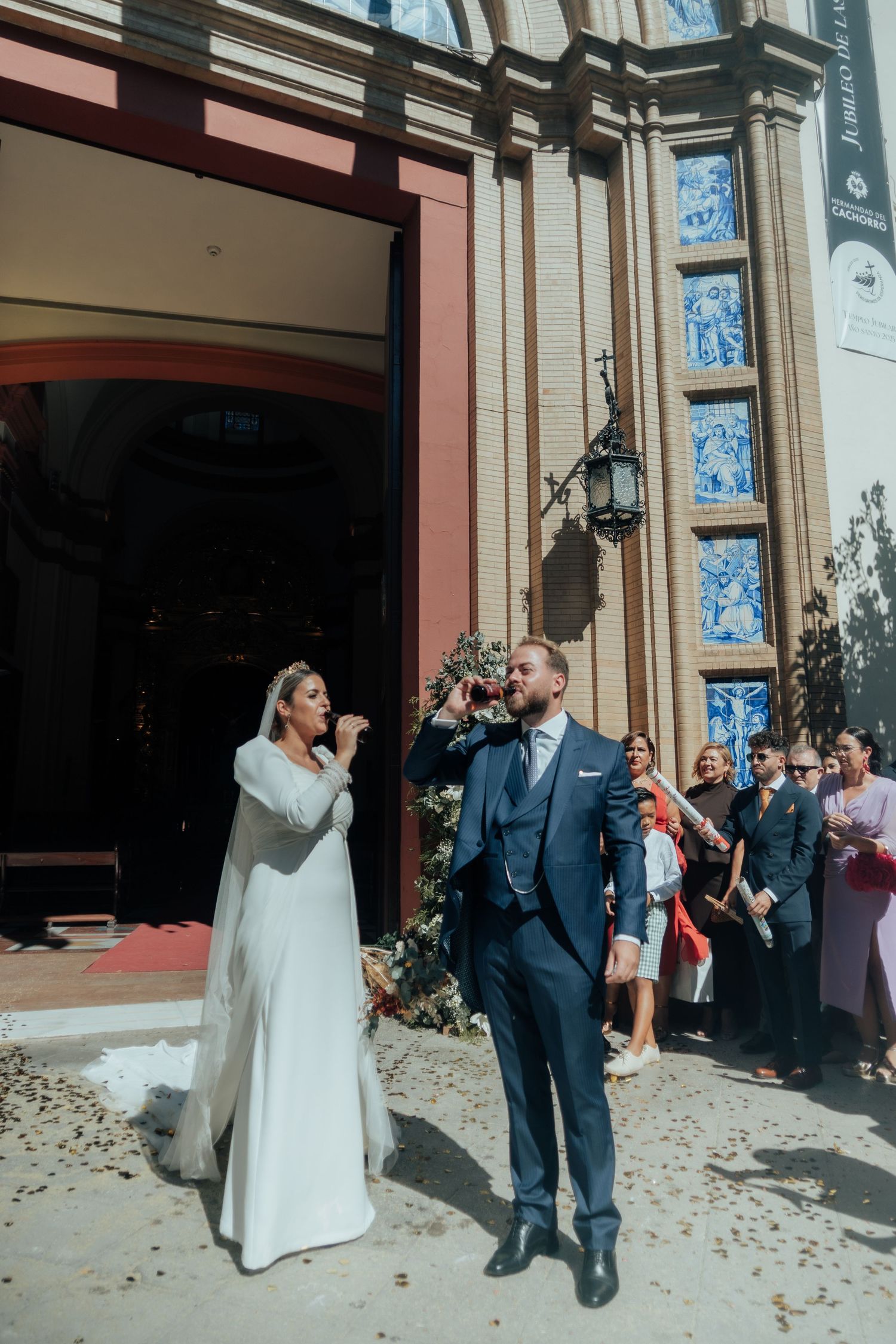 Smiling newlyweds pause outside church with blue tile panels as guests celebrate around them.