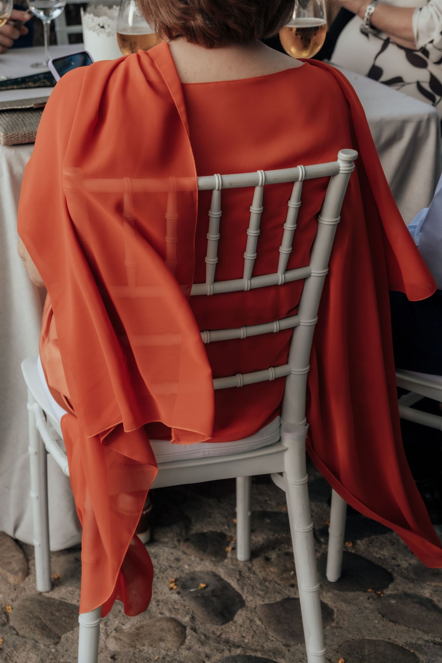 Guest in elegant orange-red flowing dress seated on white Chiavari chair at outdoor wedding reception.