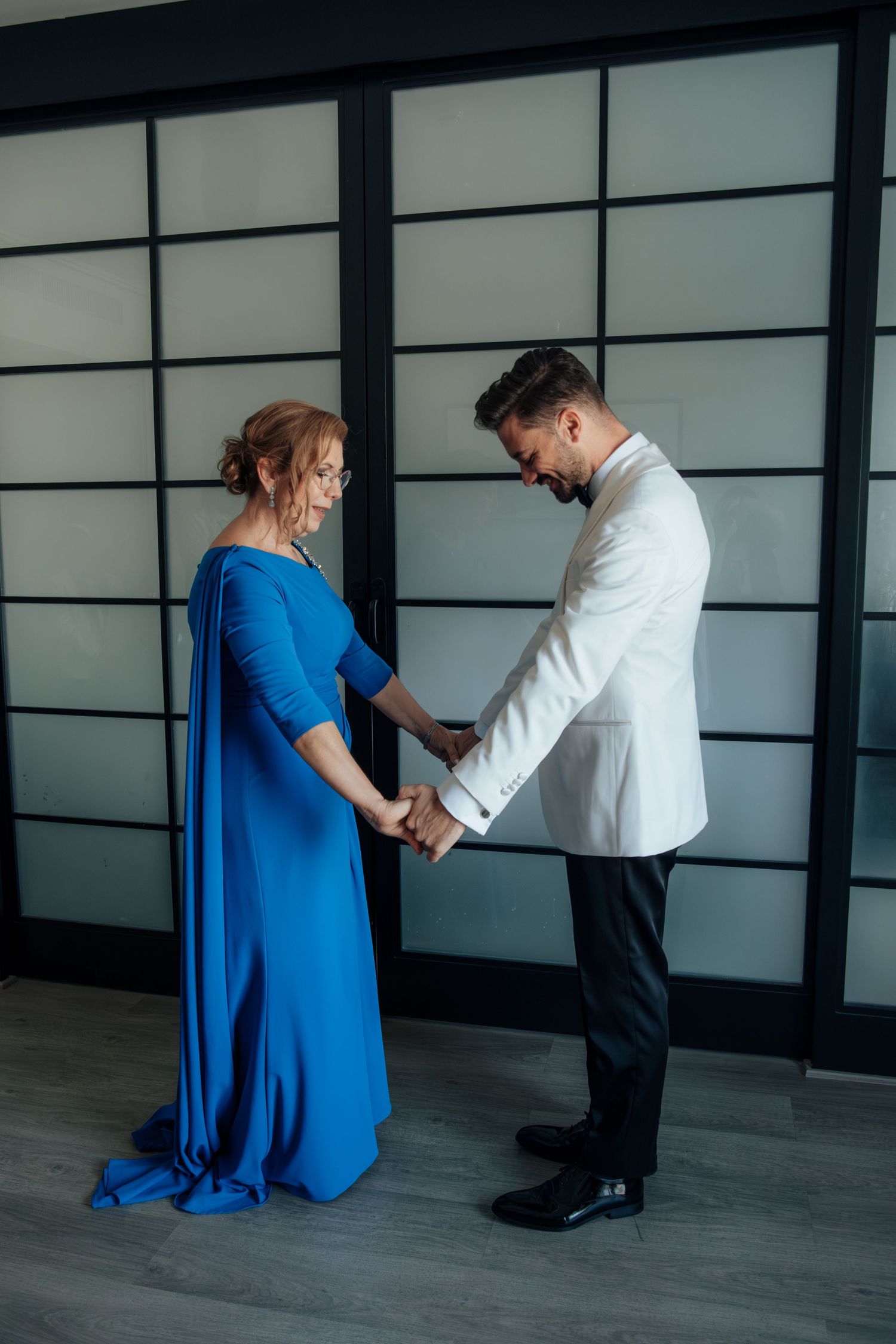 A groom in a white jacket tenderly holds hands with a woman in an elegant blue gown indoors.
