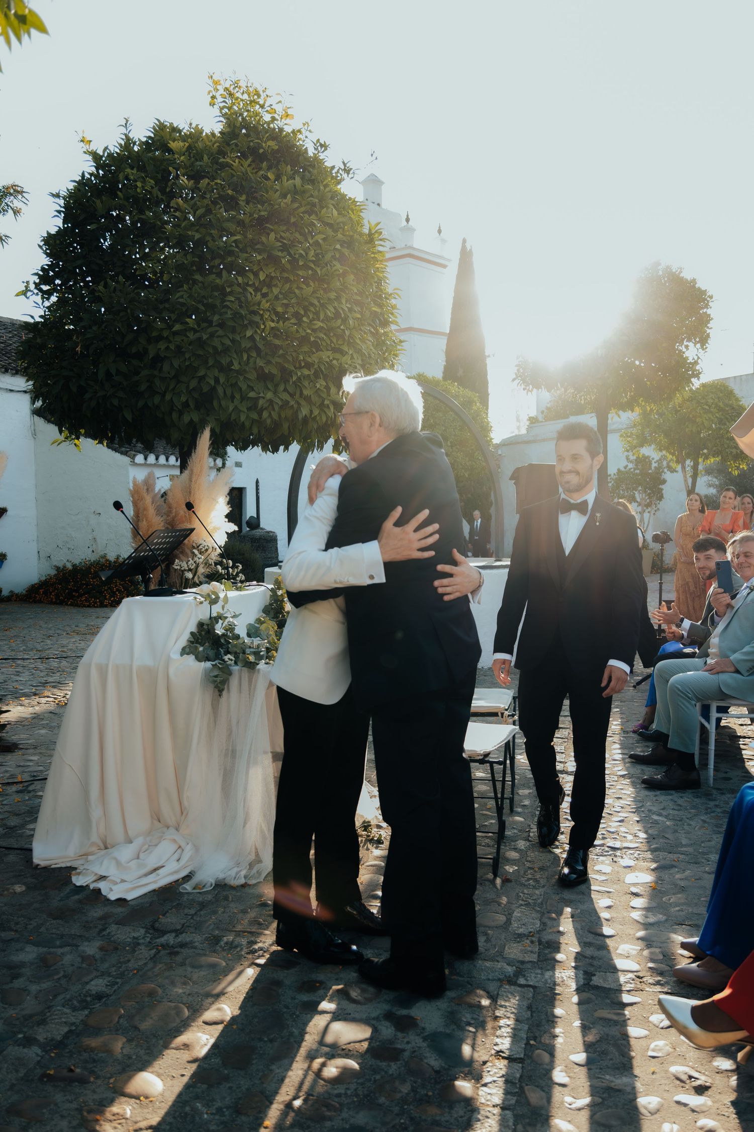 Two grooms embrace at outdoor wedding ceremony with sunlight streaming through trees on cobblestone venue.