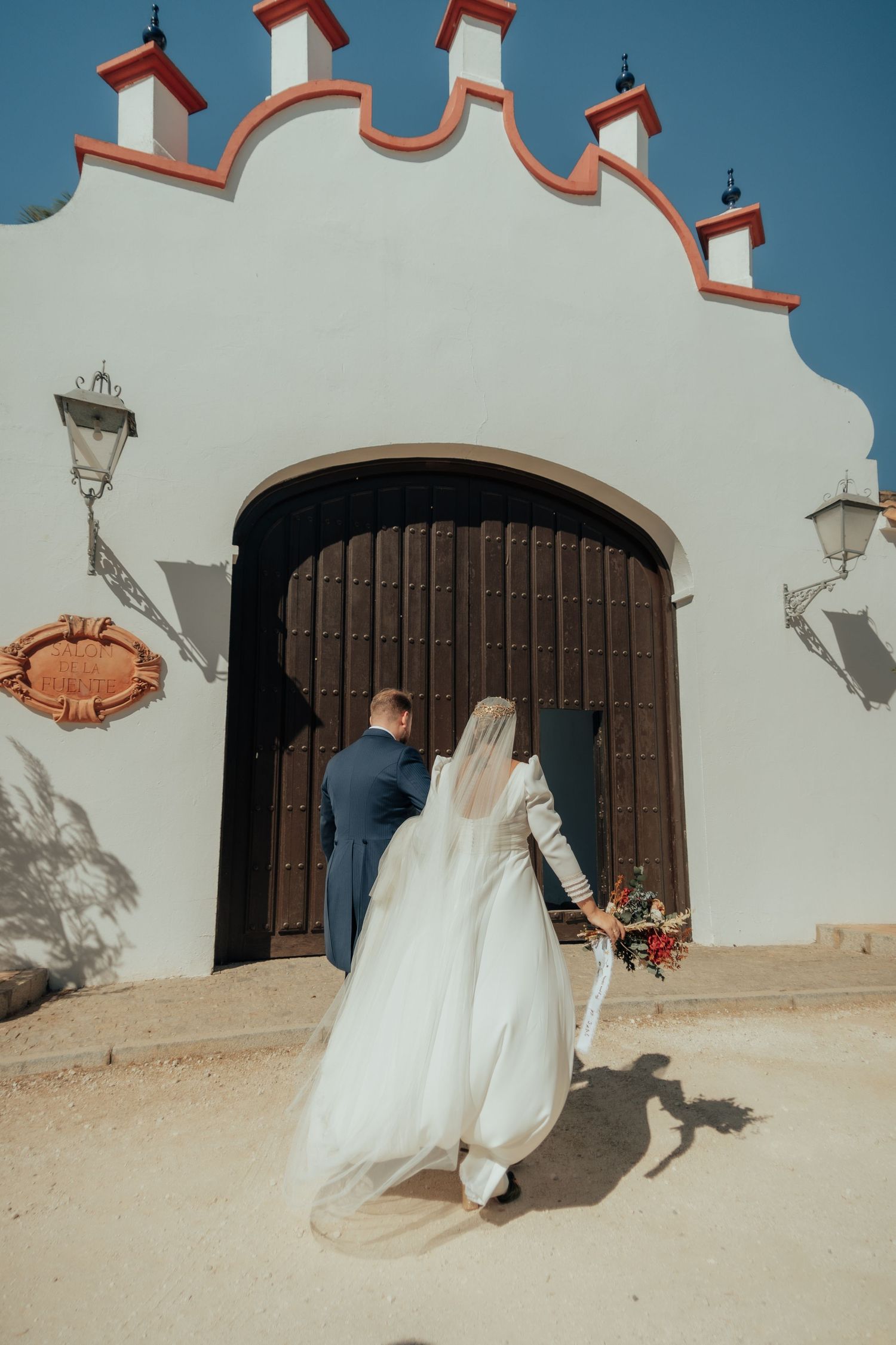 Bride in white gown and veil walks with groom toward ornate wooden doors of white Spanish chapel.