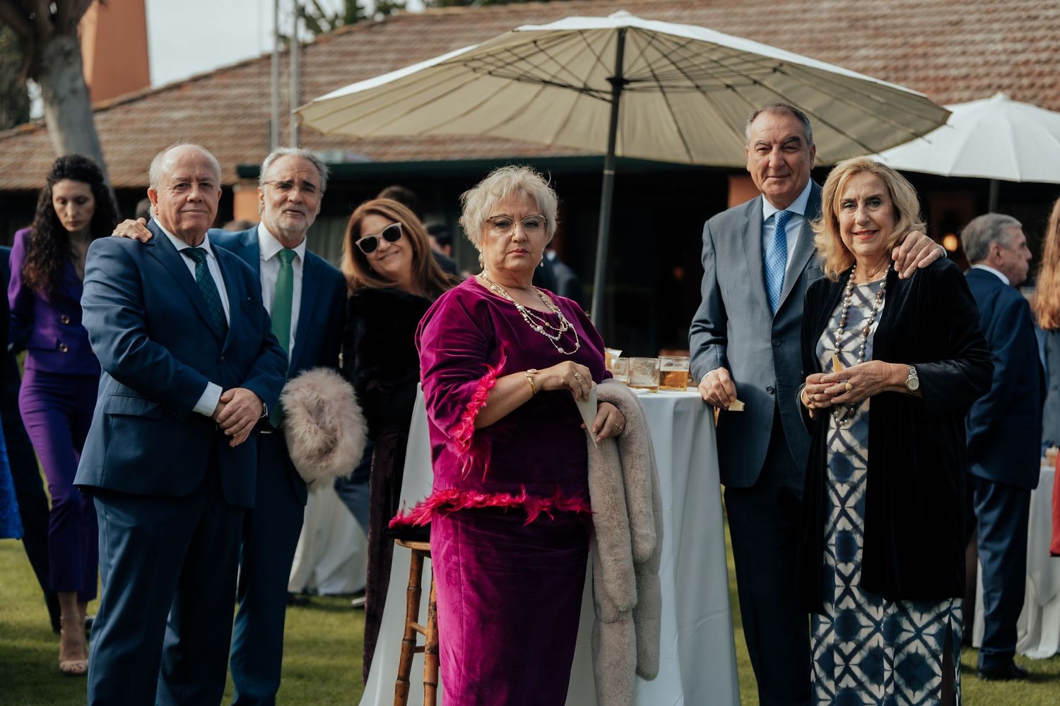 Group of six formally dressed wedding guests pose together at an outdoor cocktail reception with umbrella.