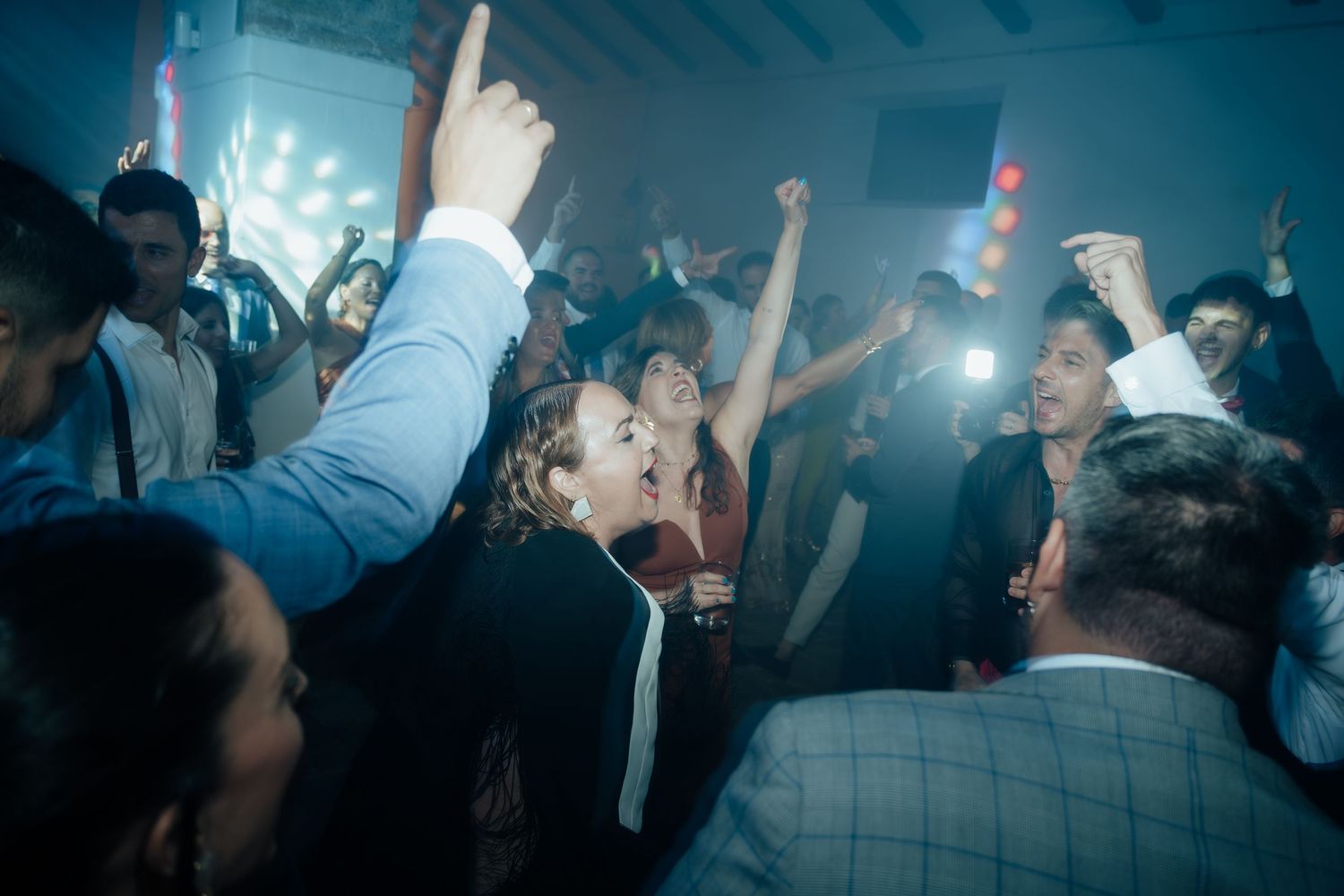 Lively wedding reception dance floor with guests celebrating, kissing, and raising hands under bright party lights.