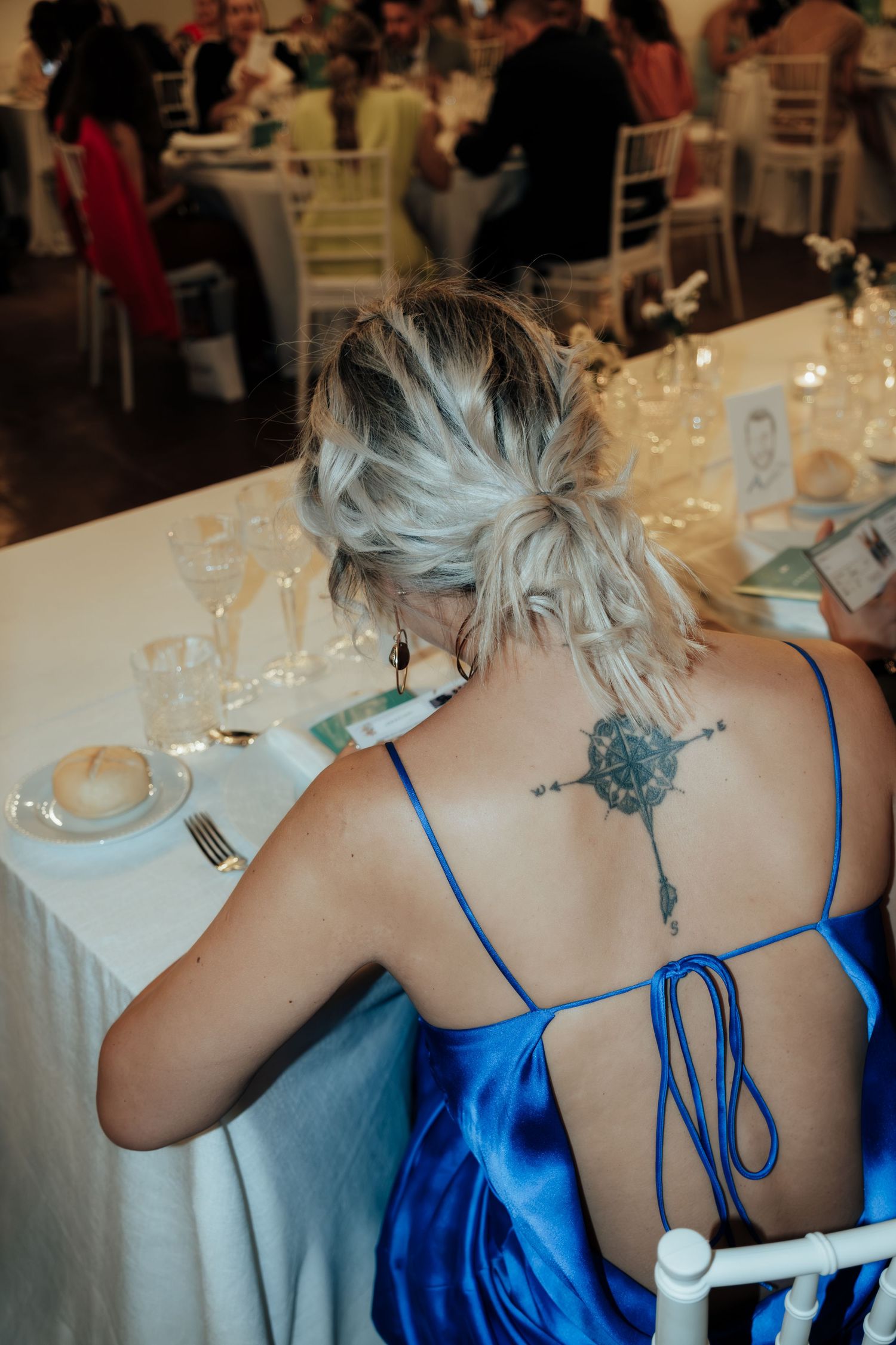 Back view of woman in blue satin dress with compass tattoo seated at a formal wedding reception table.