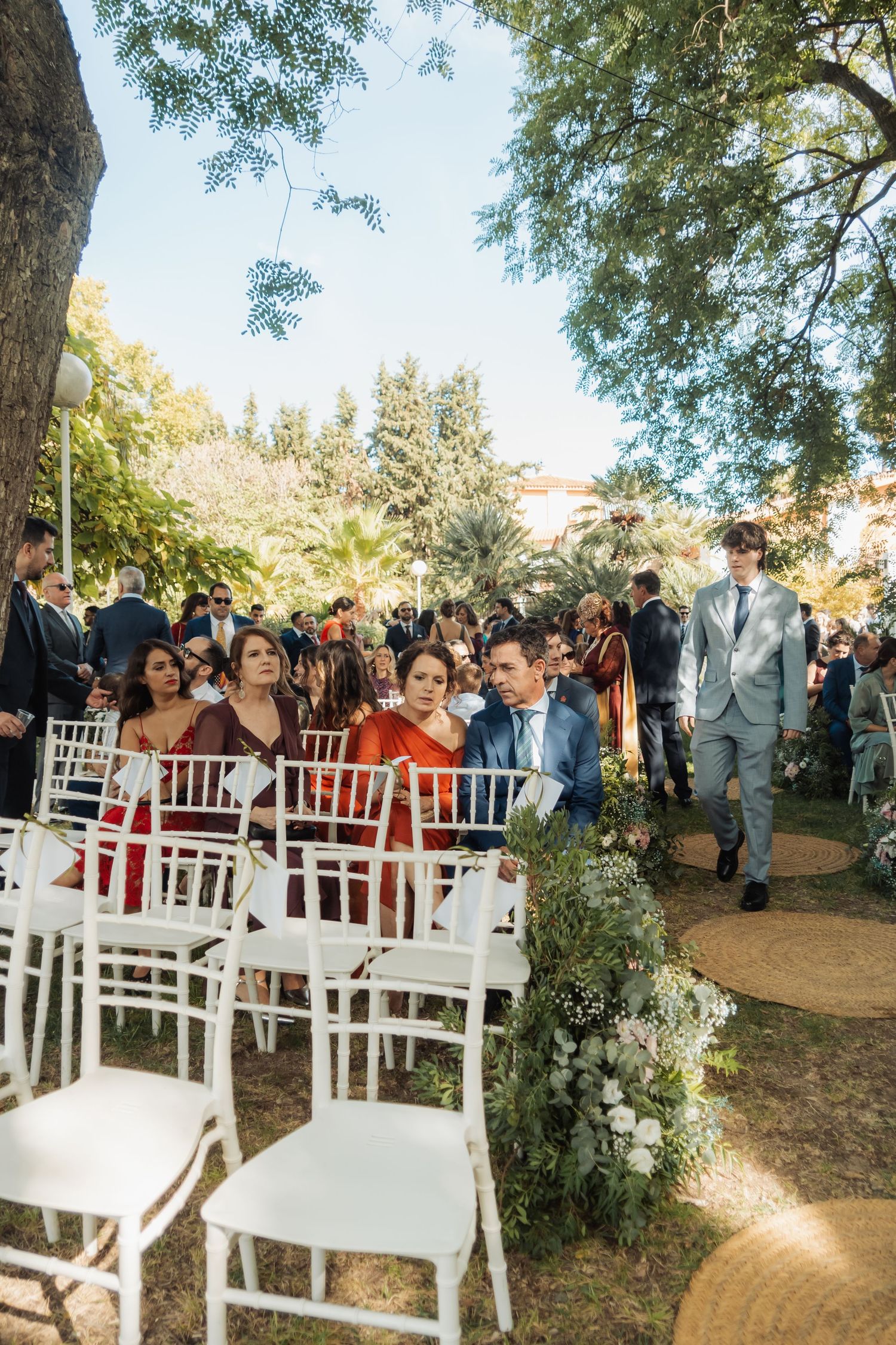 Groom in gray suit walks down aisle past seated guests at elegant outdoor garden wedding ceremony.