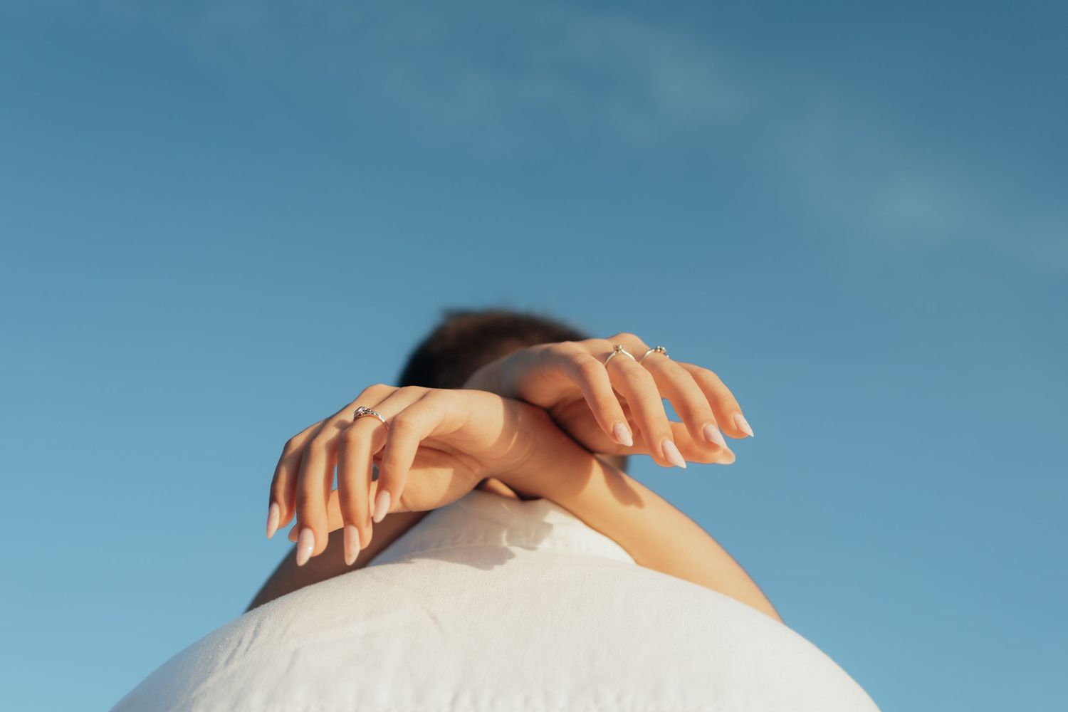 Woman in white dress crossing arms over head against blue sky, showcasing manicured nails.