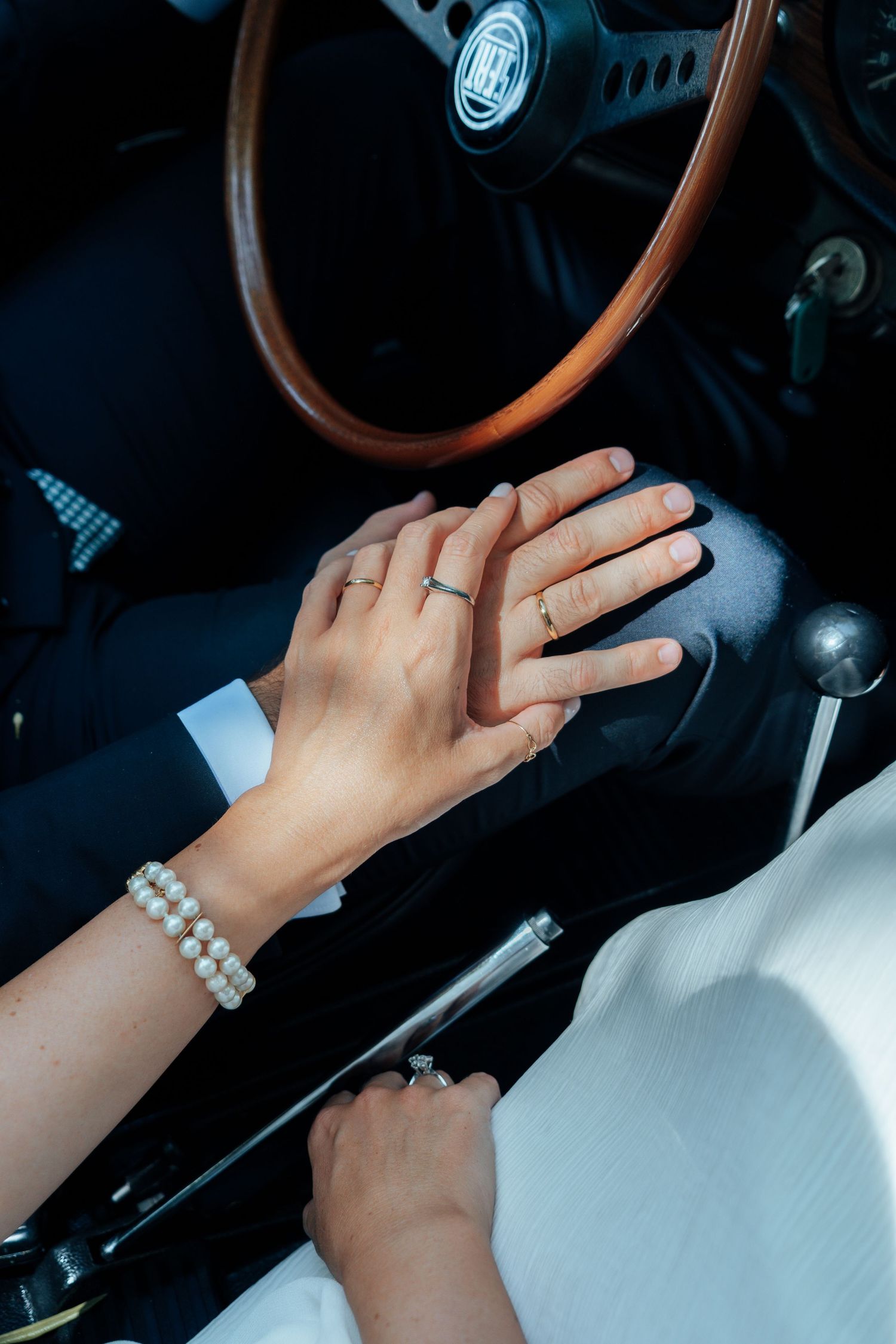Close-up of bride and groom holding hands inside a vintage car, both wearing wedding rings.