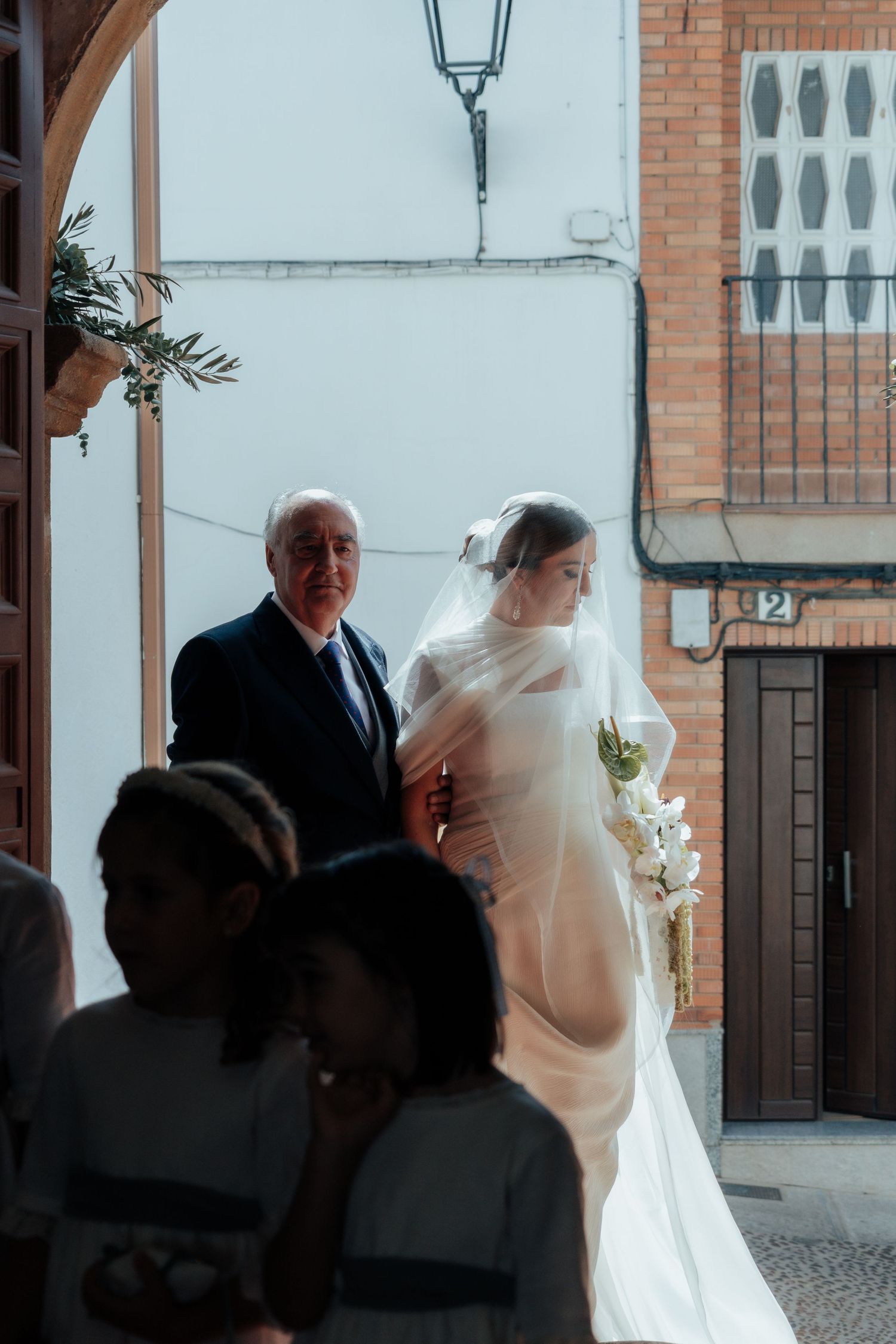 Bride in white veil holding floral bouquet walks with older man outside brick building.