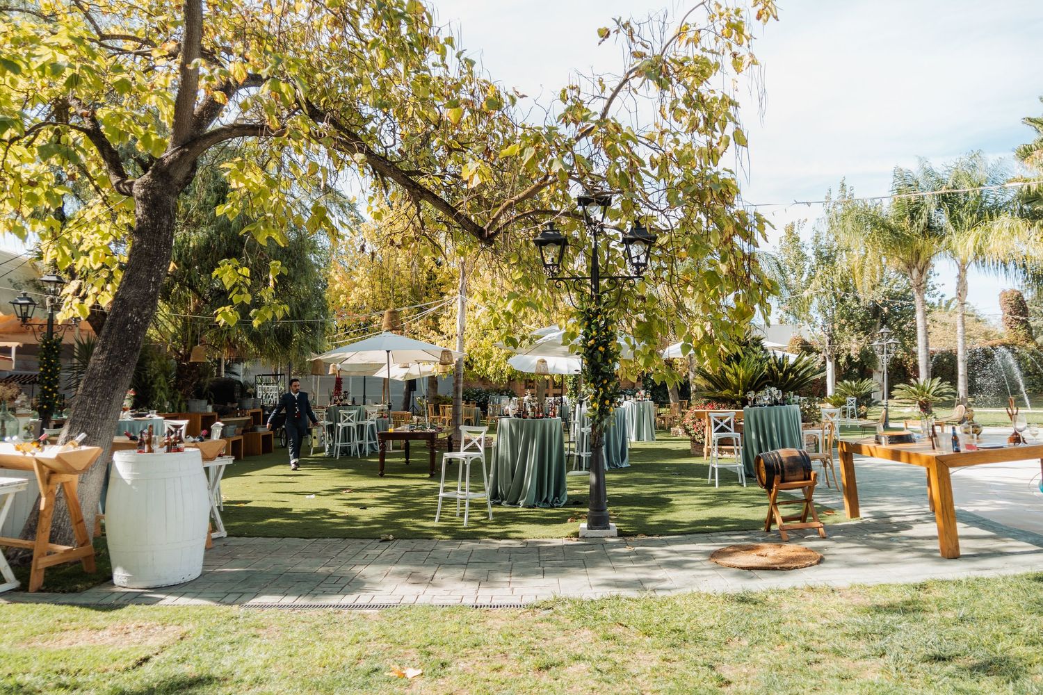 Elegant outdoor wedding reception venue with green lawn, trees, white umbrellas, and wooden furniture.