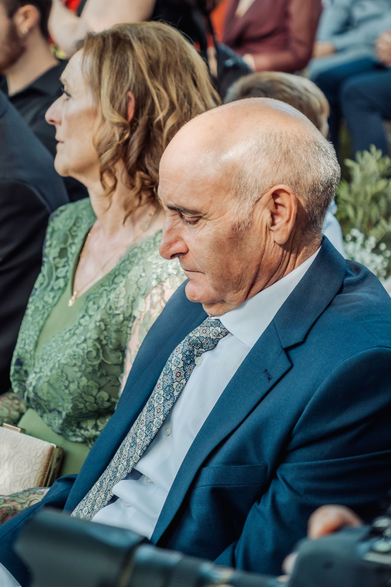 Elderly bald man in blue suit and tie seated attentively at an outdoor wedding ceremony.