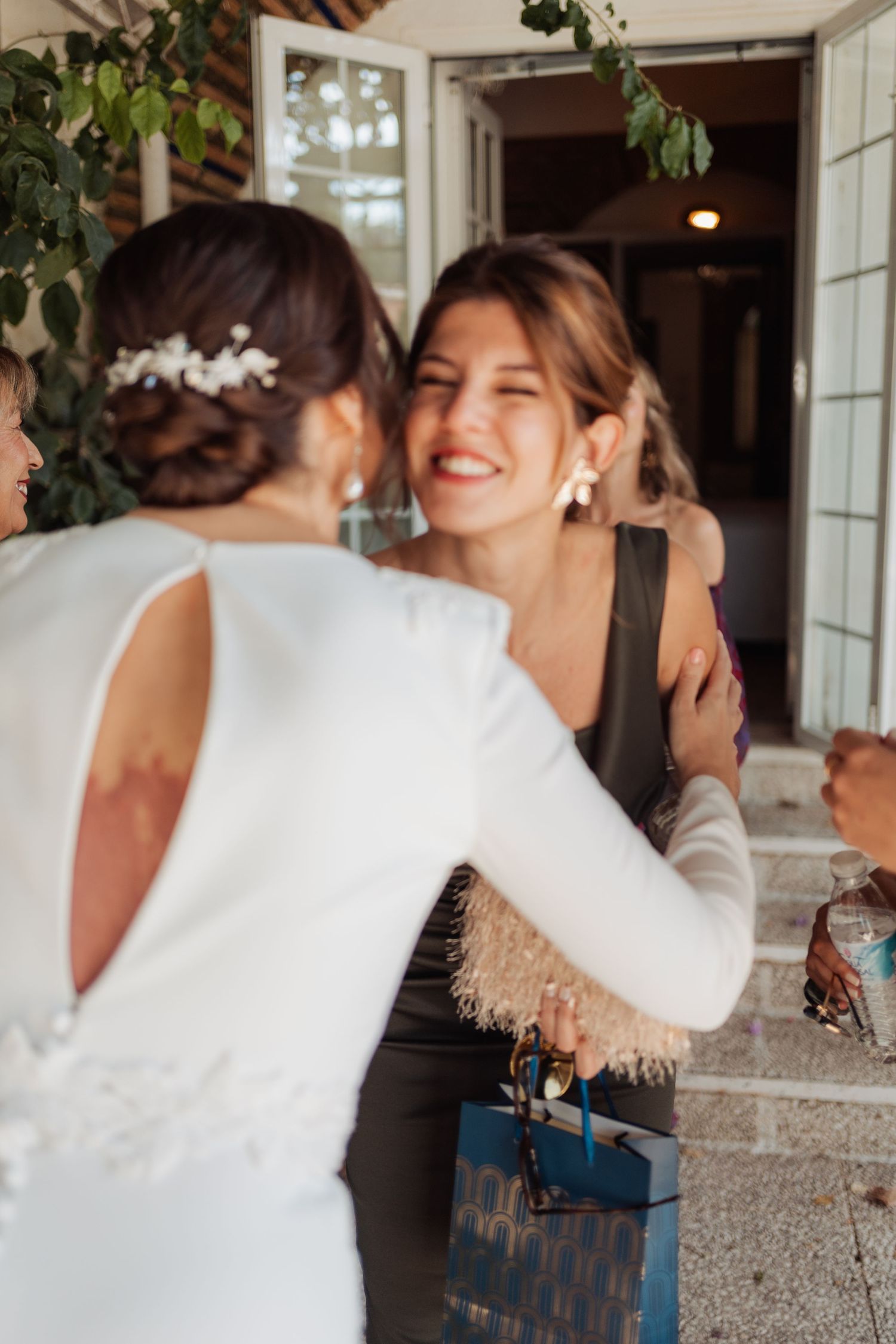 Joyful wedding guest smiles and embraces bride in white gown at outdoor wedding venue entrance.