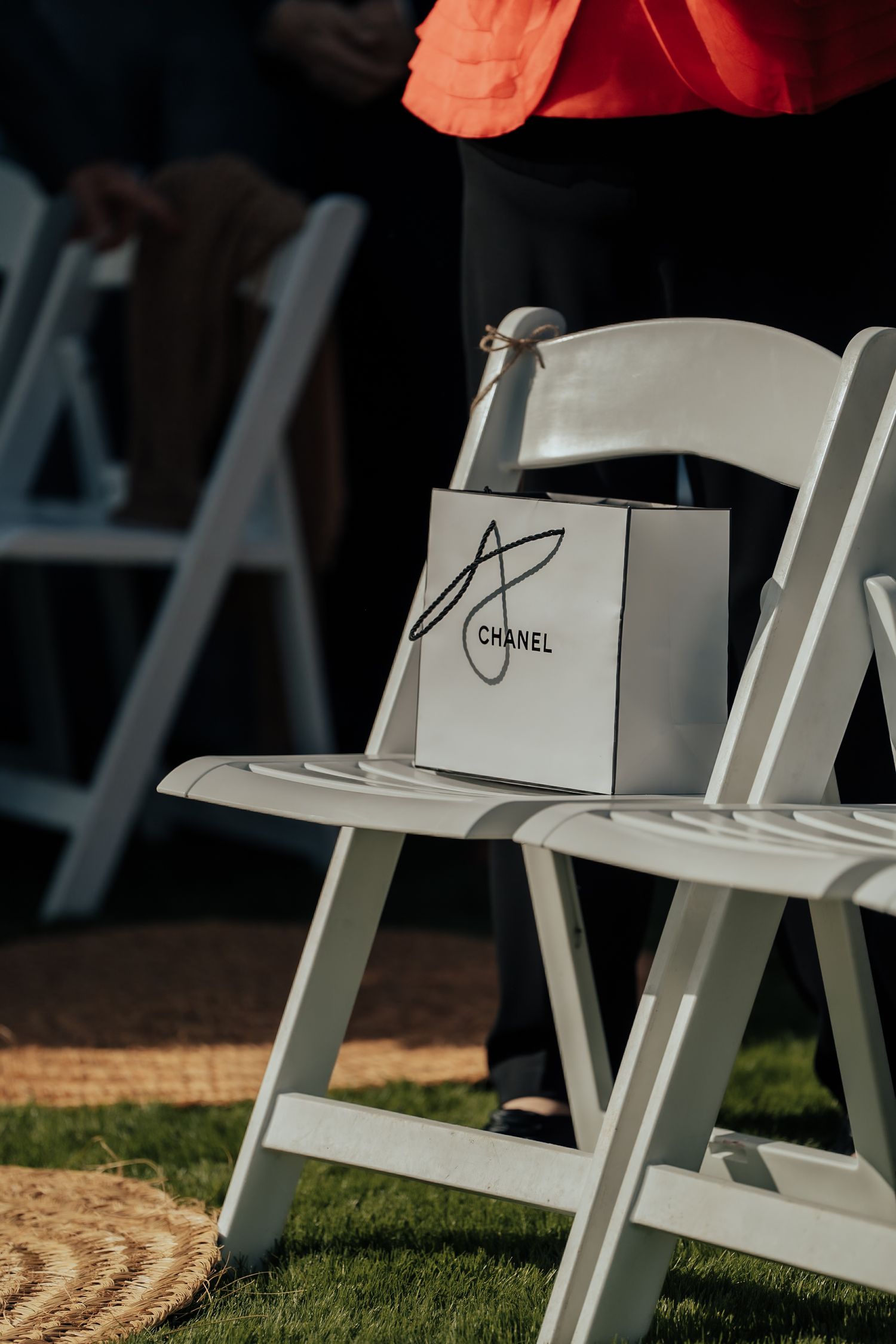 White Chanel gift bag placed on a white folding chair at an outdoor wedding ceremony setup on a sunny day.