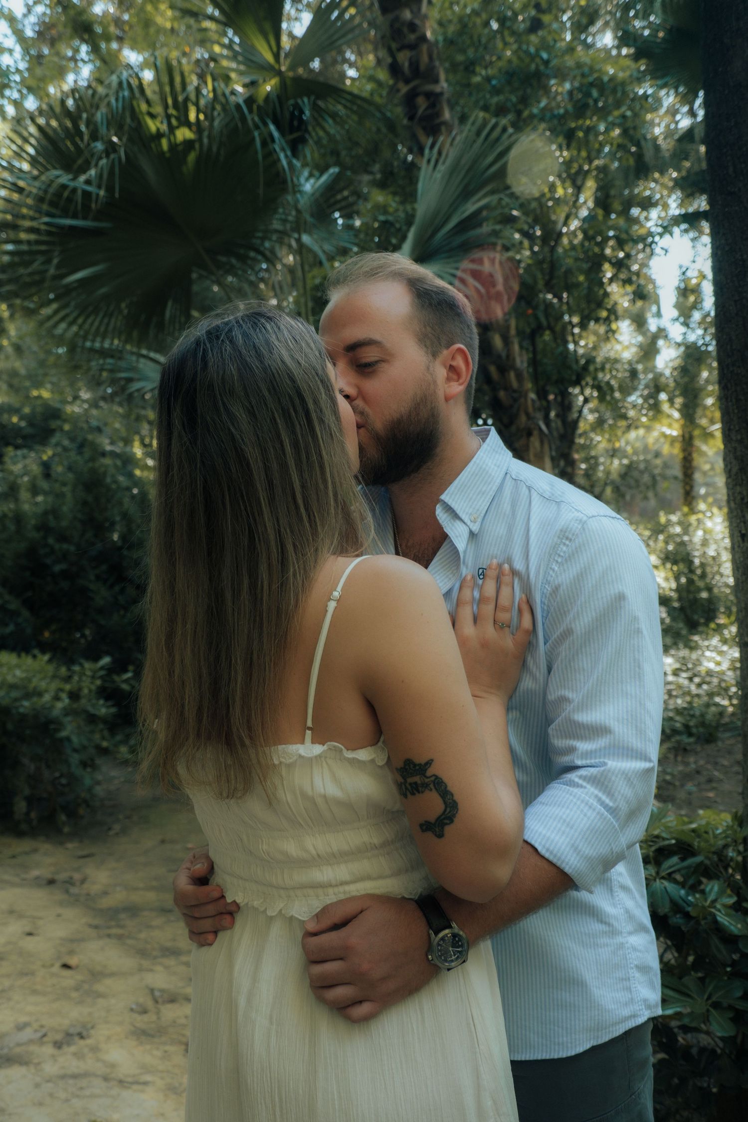 Couple sharing a romantic kiss surrounded by lush tropical greenery, woman in white dress with tattoo visible.