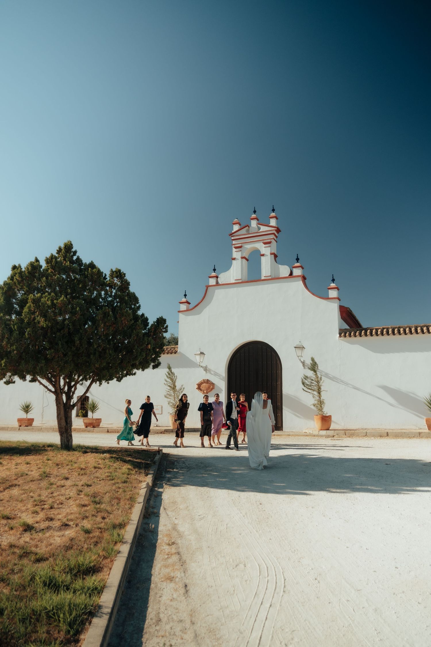 Wedding guests arriving at a white Spanish-style chapel with red accents under a clear blue sky.