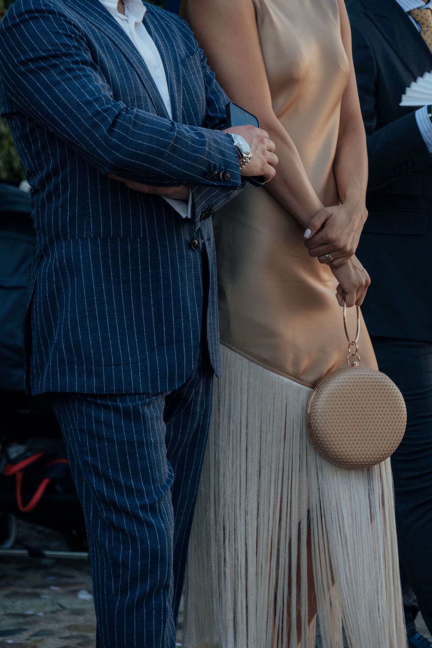 Couple in formal attire at wedding, man in navy pinstripe suit, woman in gold dress holding round bag.
