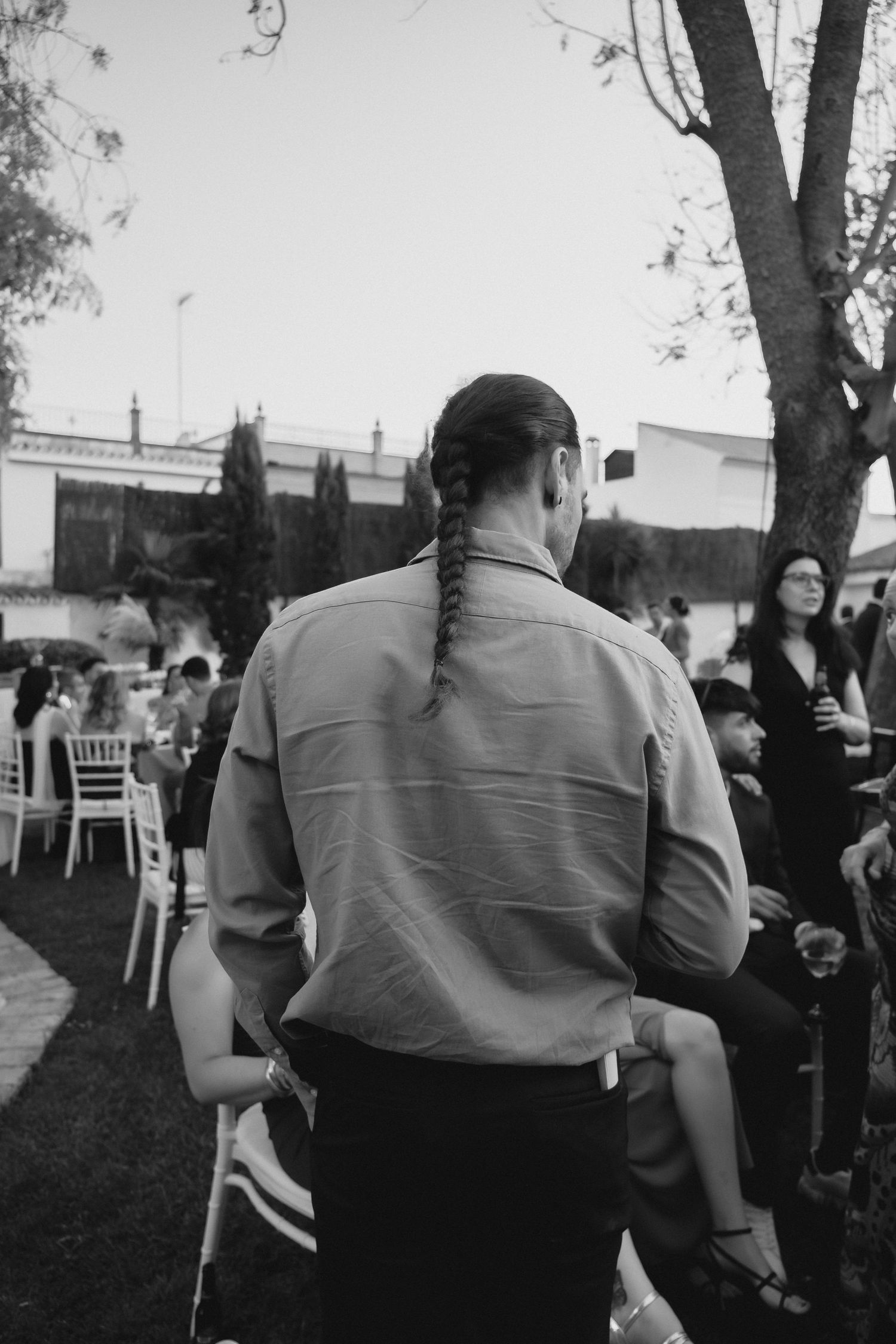 Black and white photo of man with braided hair at outdoor wedding reception.