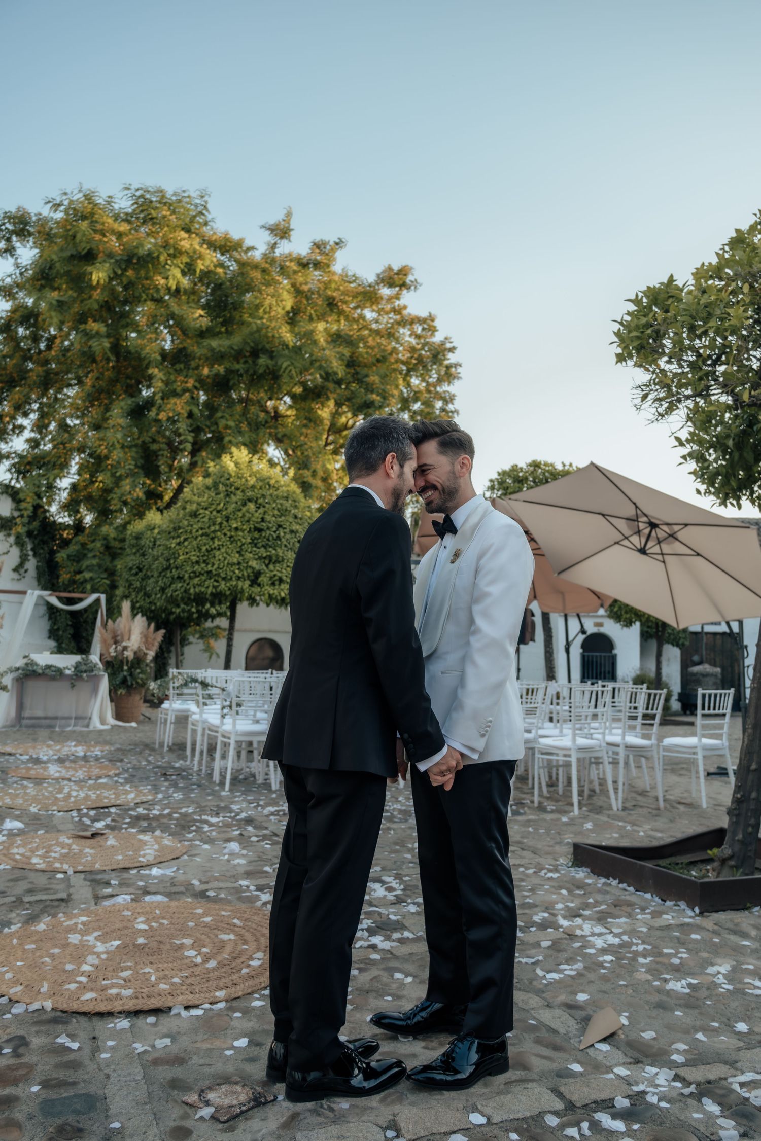 Two grooms share intimate moment holding hands in empty outdoor wedding venue after ceremony.