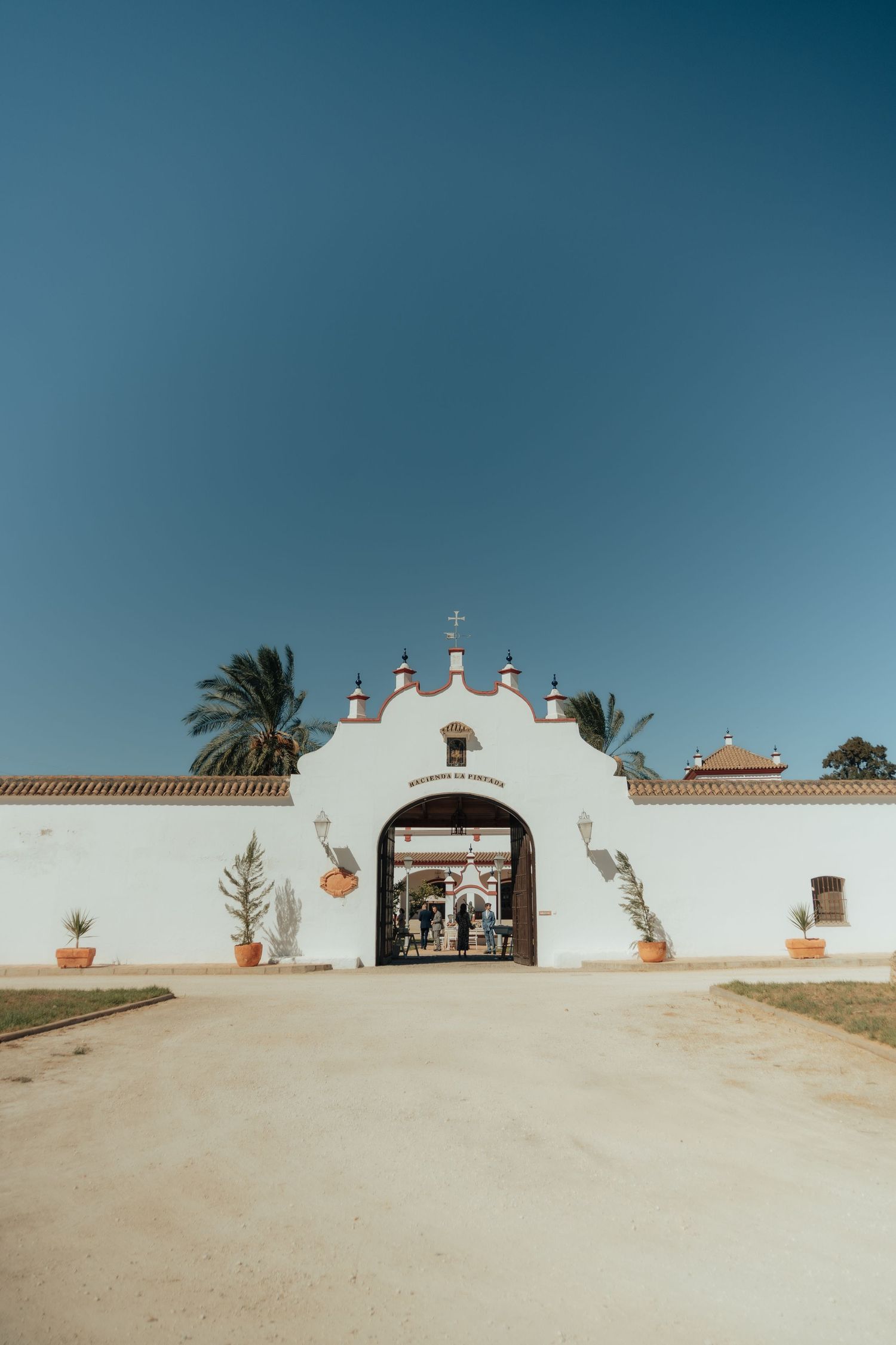 White Spanish hacienda wedding venue entrance with cross, arch, and palm trees under blue sky.