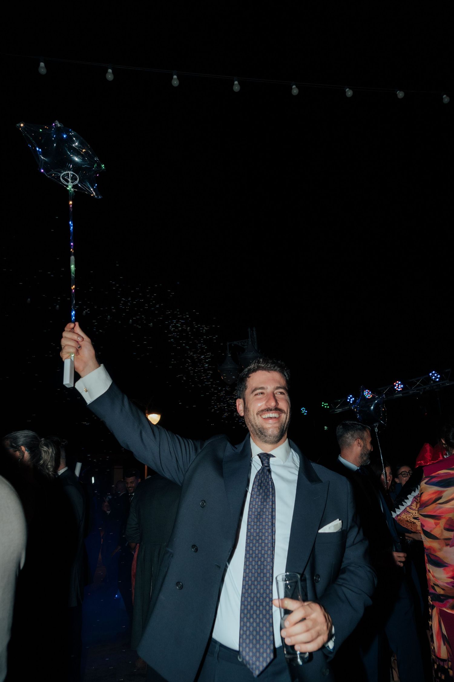 Joyful man in suit raises a glowing bubble wand at a festive outdoor wedding reception.