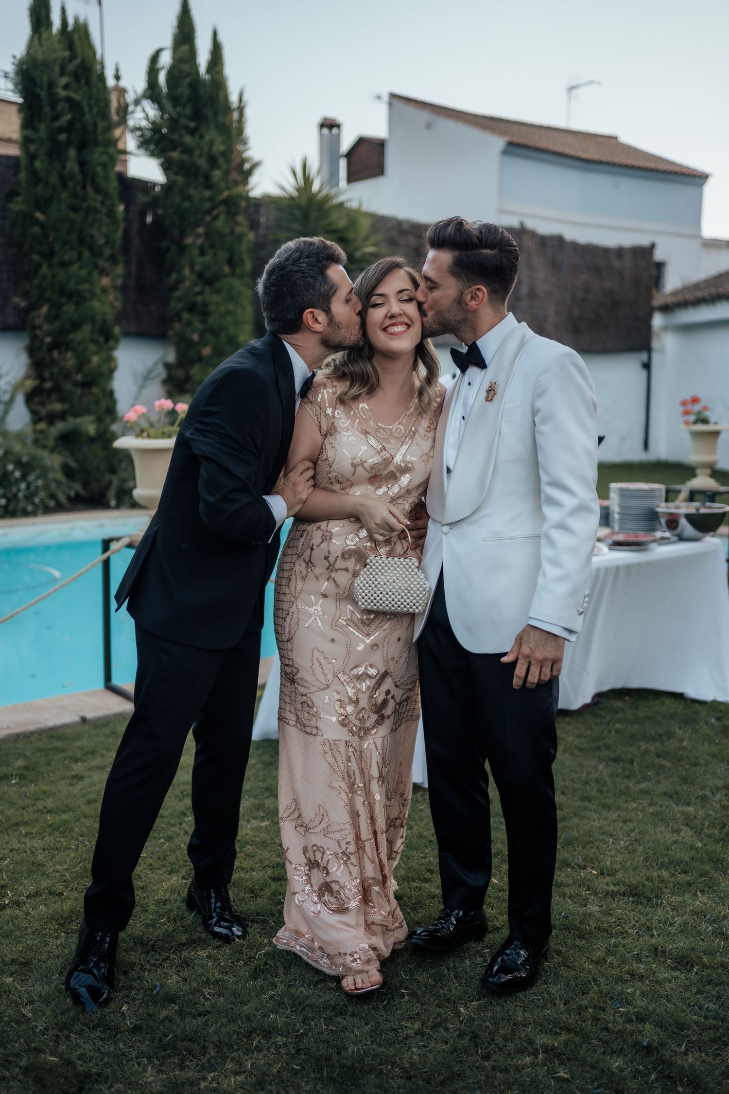 Two grooms in tuxedos kissing a woman in gold sequin gown on cheeks at outdoor poolside wedding reception.