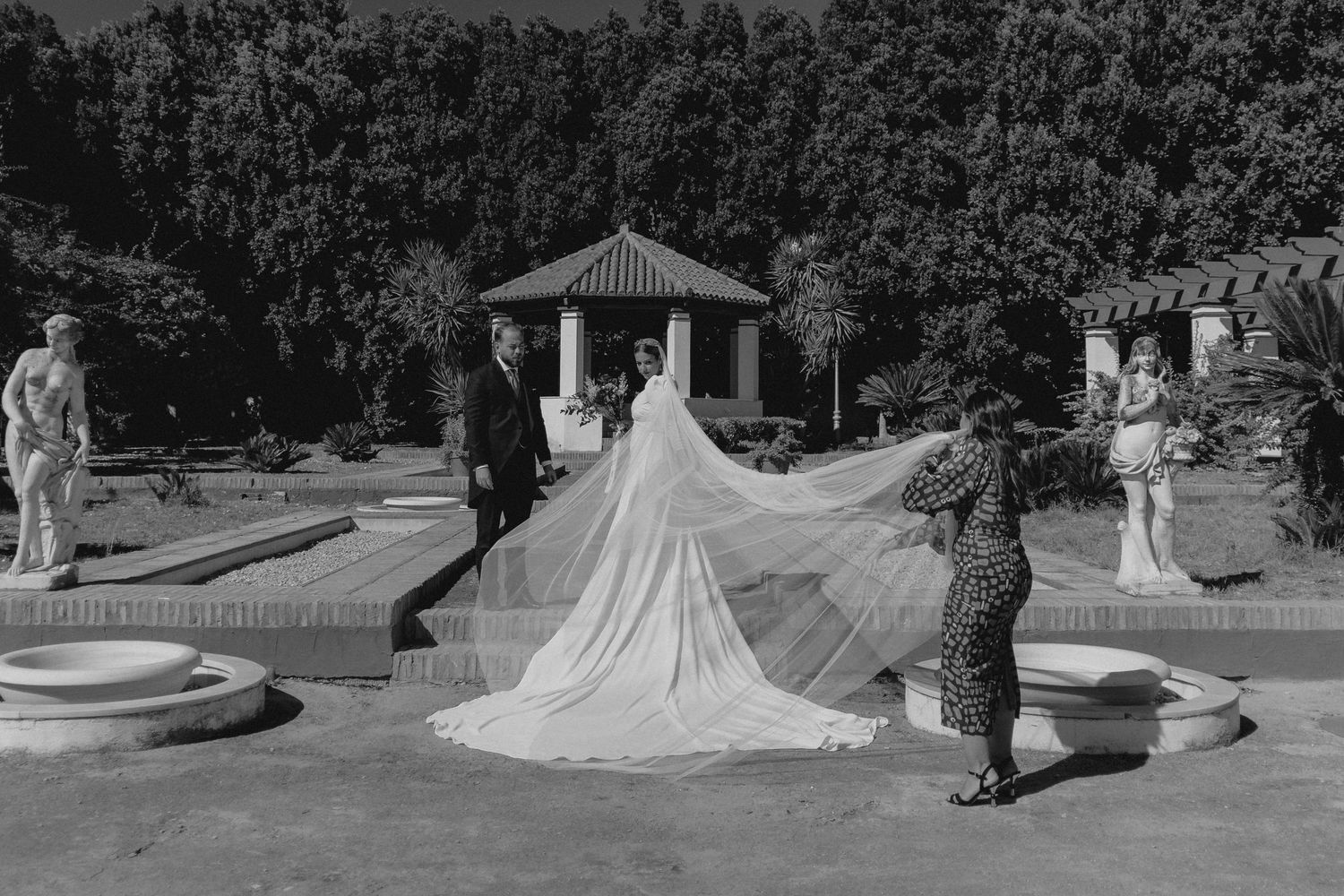 Black and white photo of bride with long veil being adjusted by attendants in a formal garden.
