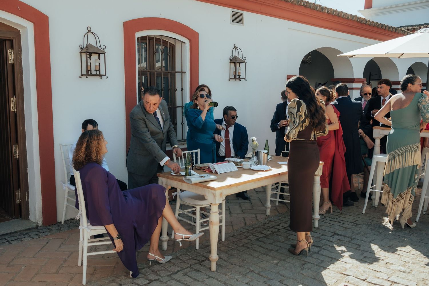 Wedding guests mingling outside white venue with red trim, registration table in foreground.