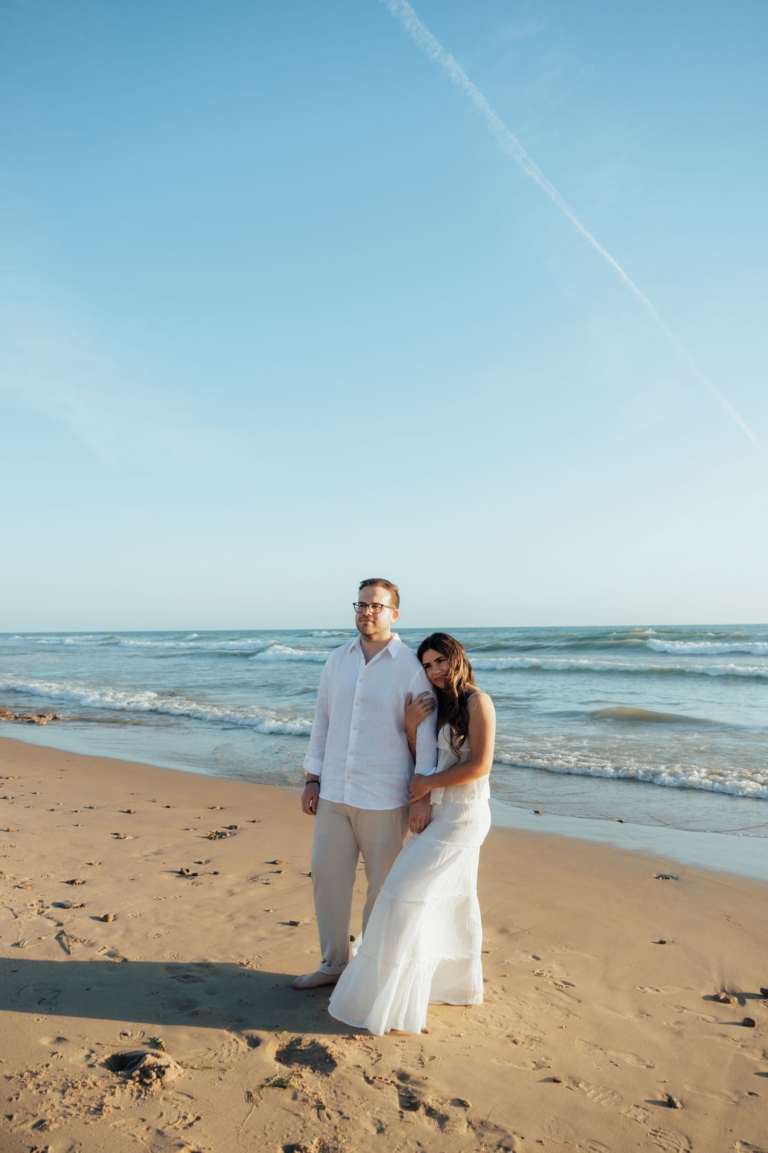 Couple in white attire posing on a sunny beach with ocean waves and blue sky backdrop.
