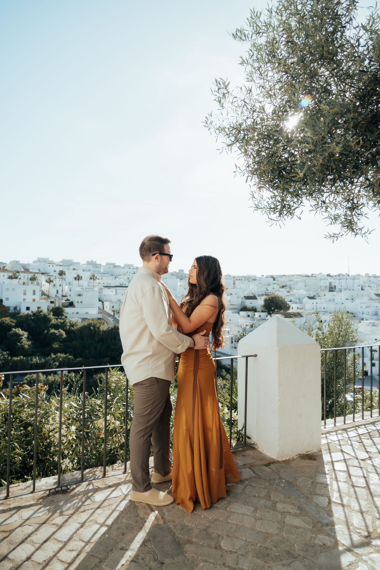 Couple embracing on a scenic terrace overlooking a white-washed Mediterranean village, woman in golden dress.