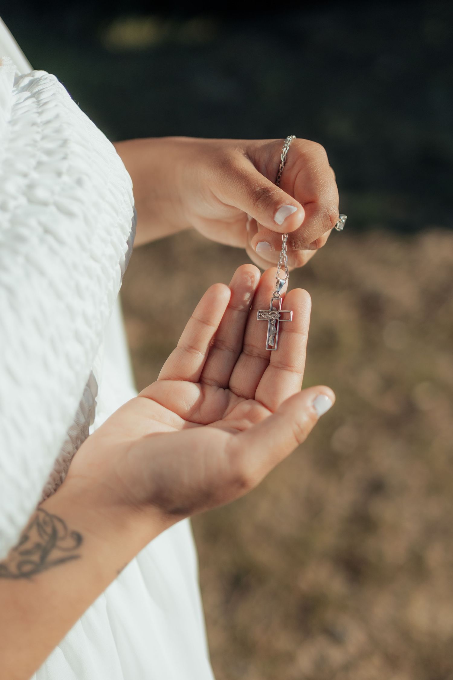 Hands holding a silver cross necklace with rosary beads, wearing white dress with butterfly tattoo visible.
