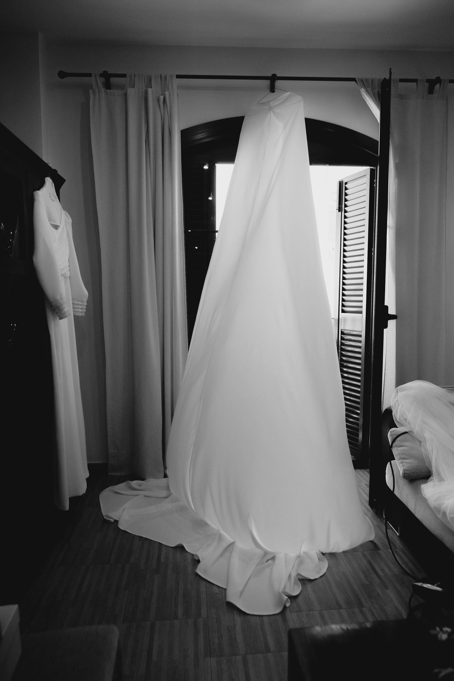 Black and white photo of elegant wedding gown hanging near arched window in bridal suite.