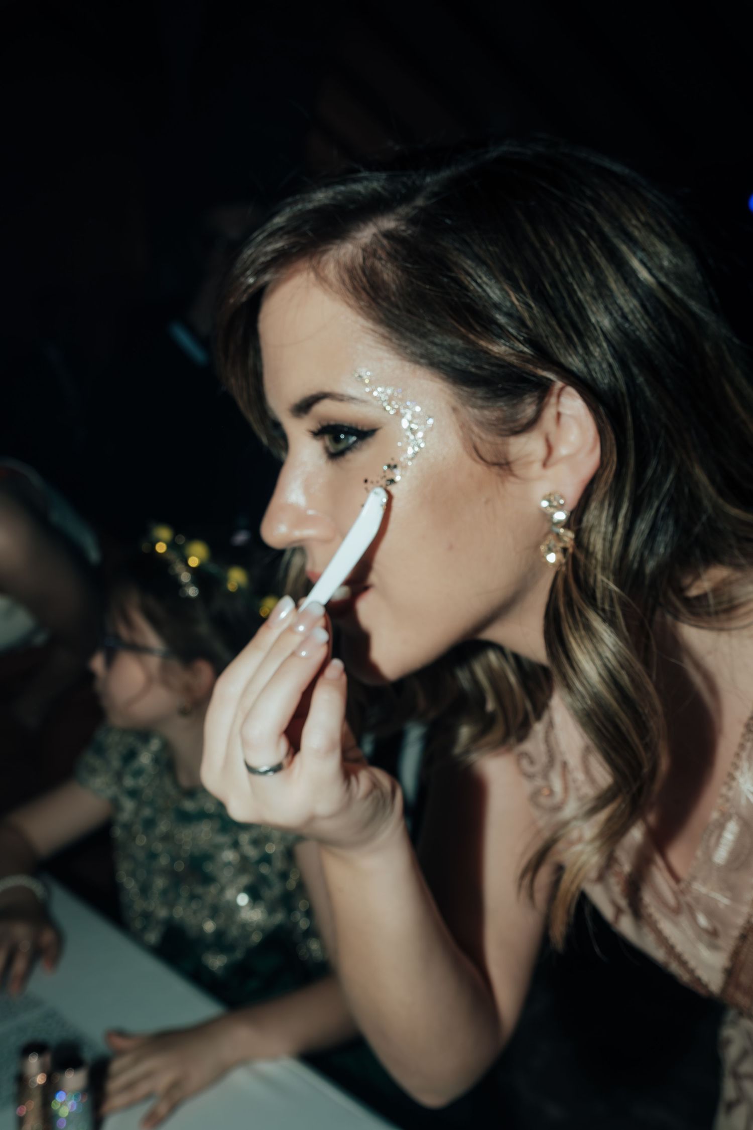 A young woman with glitter face makeup applies lipstick at a glamorous masquerade party event.