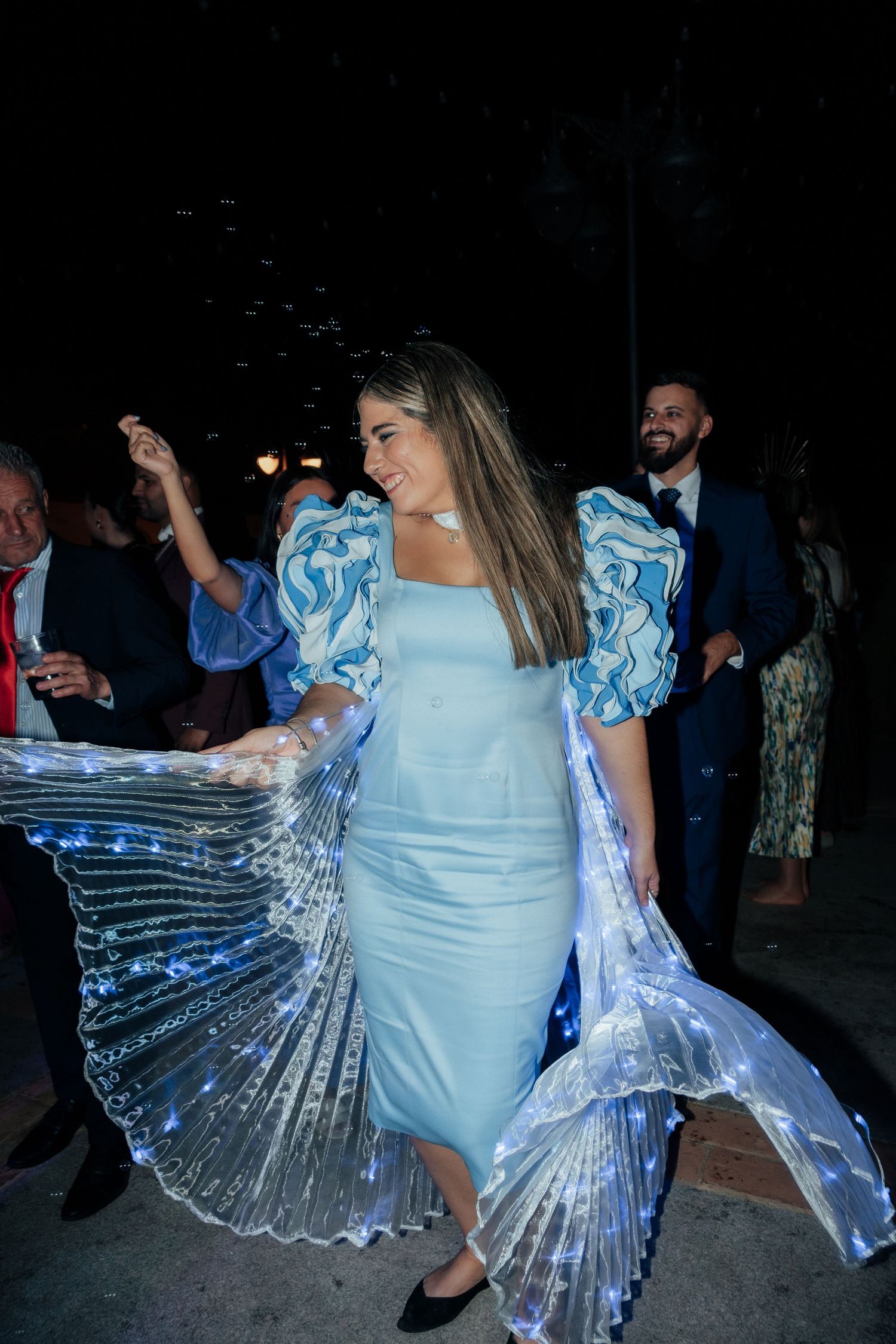Woman in light blue dress with illuminated LED wings dancing at night celebration.
