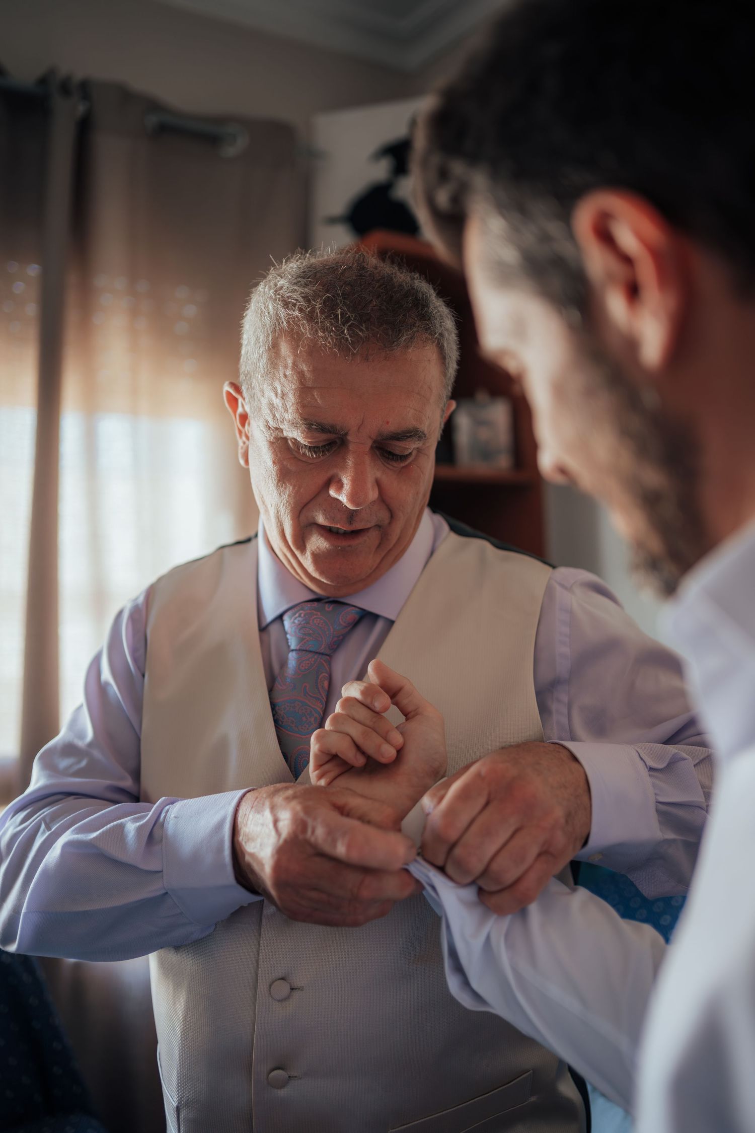 An older man in a white vest and lavender shirt gets his cufflinks fastened by another man during wedding preparations.