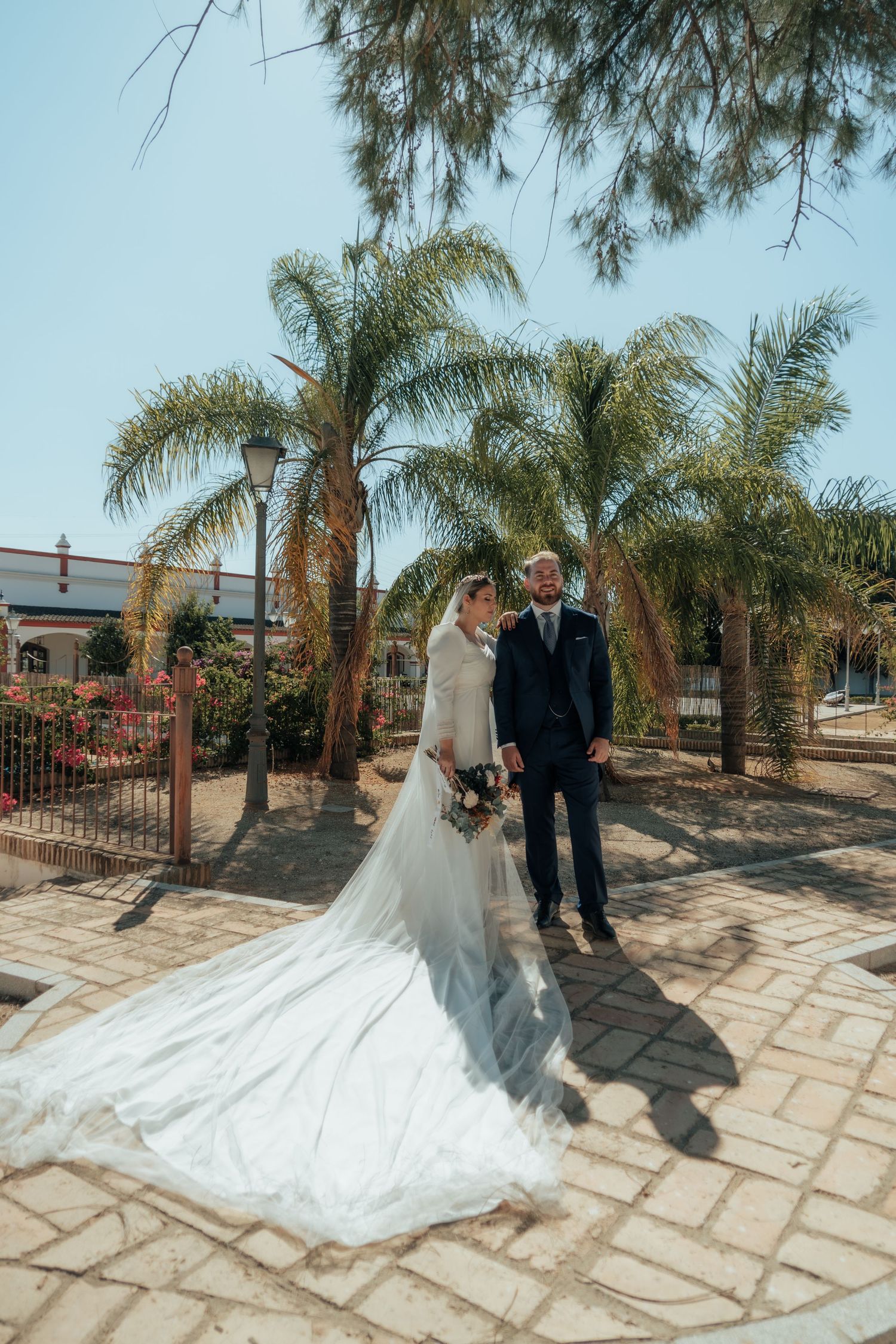 Bride and groom stand together on brick path surrounded by palm trees under a bright blue sky.