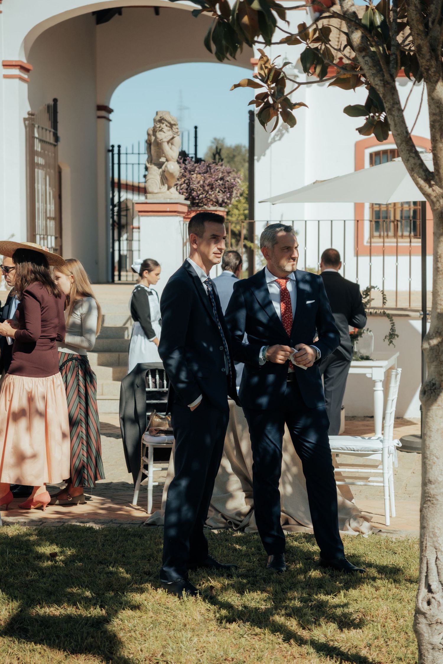 Two men in dark suits walking on grass at outdoor wedding venue with arch.