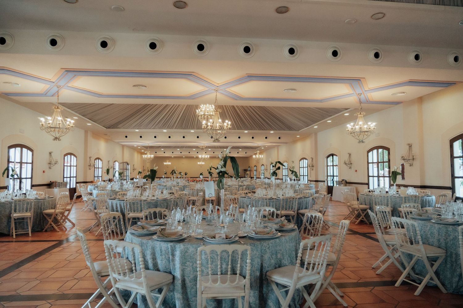 Spacious wedding reception hall with round tables, white chairs, and ornate chandeliers.