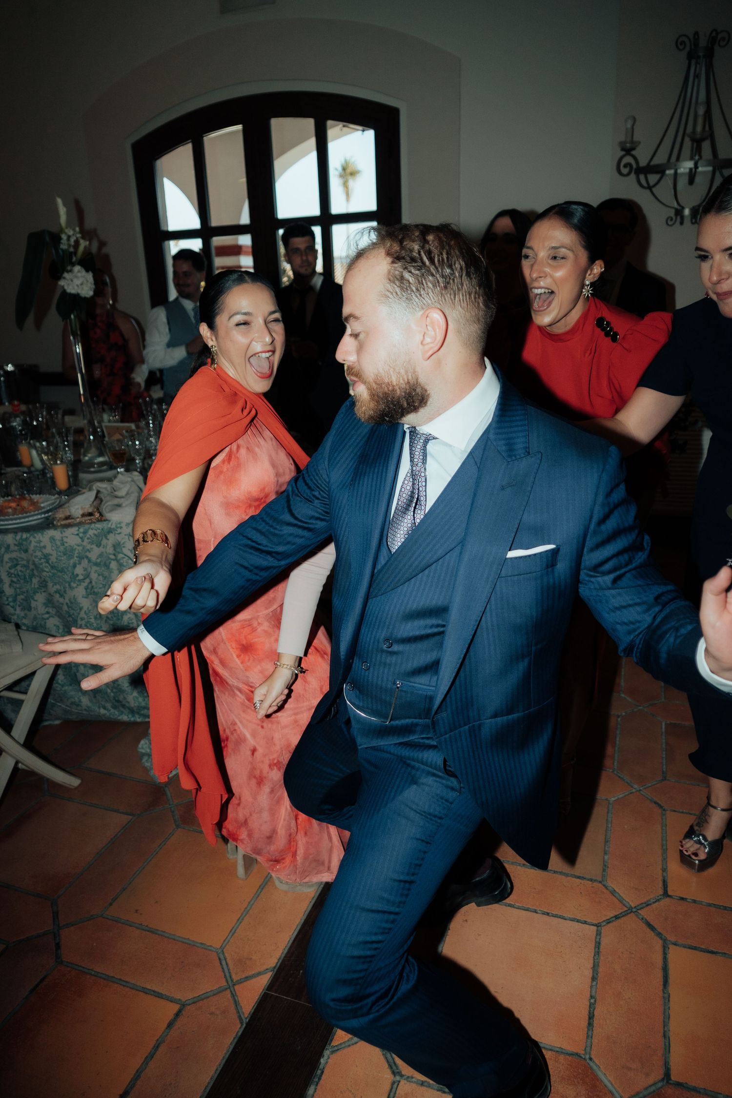 Bearded groom in navy suit dances joyfully with guests in red dresses at wedding reception.