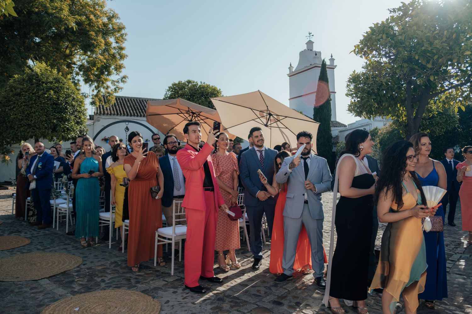 Elegantly dressed wedding guests gather outdoors in vibrant colorful attire at a sunny celebration.
