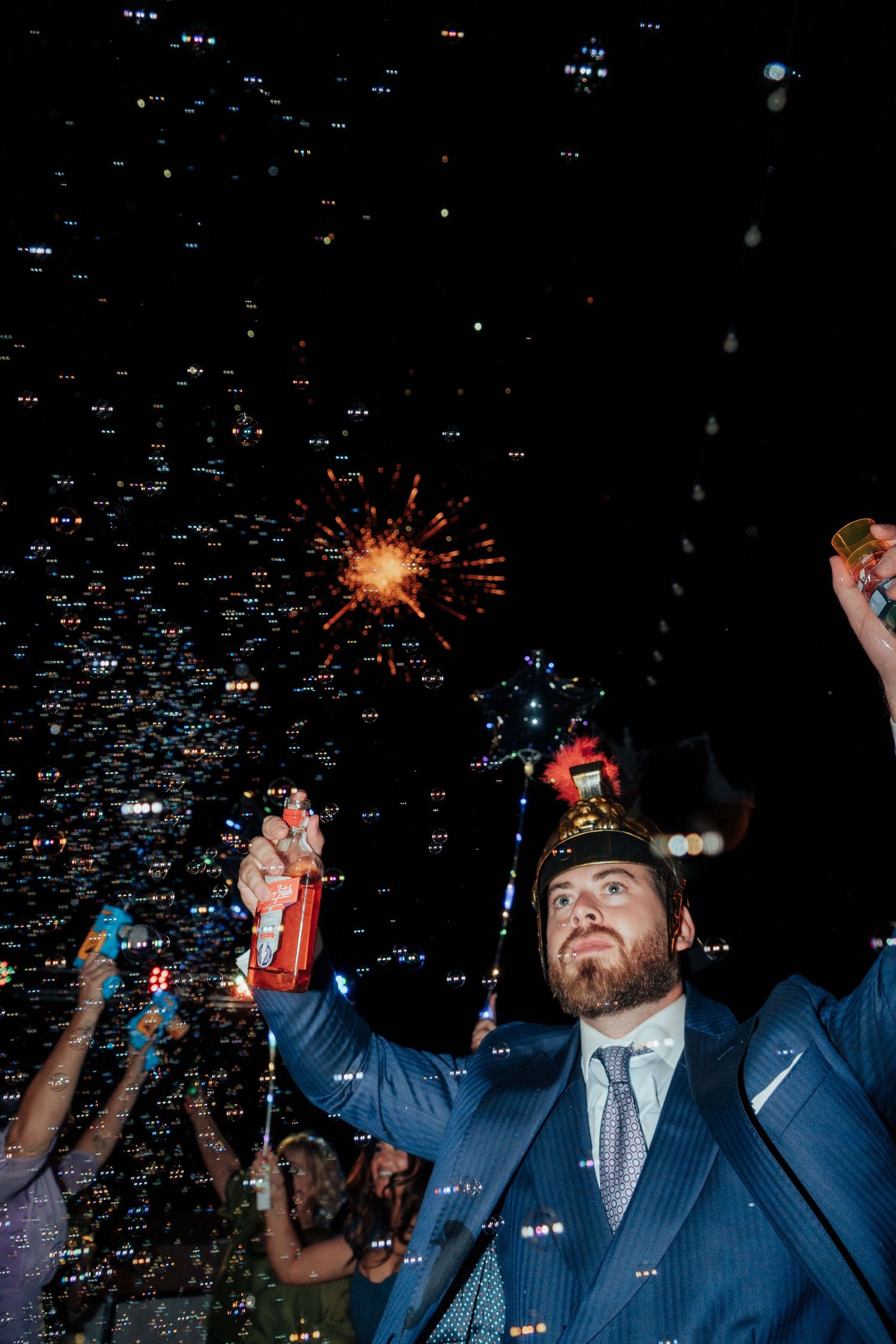 A bearded man in a blue suit celebrates at night holding a sparkler and confetti cannon at an outdoor party.